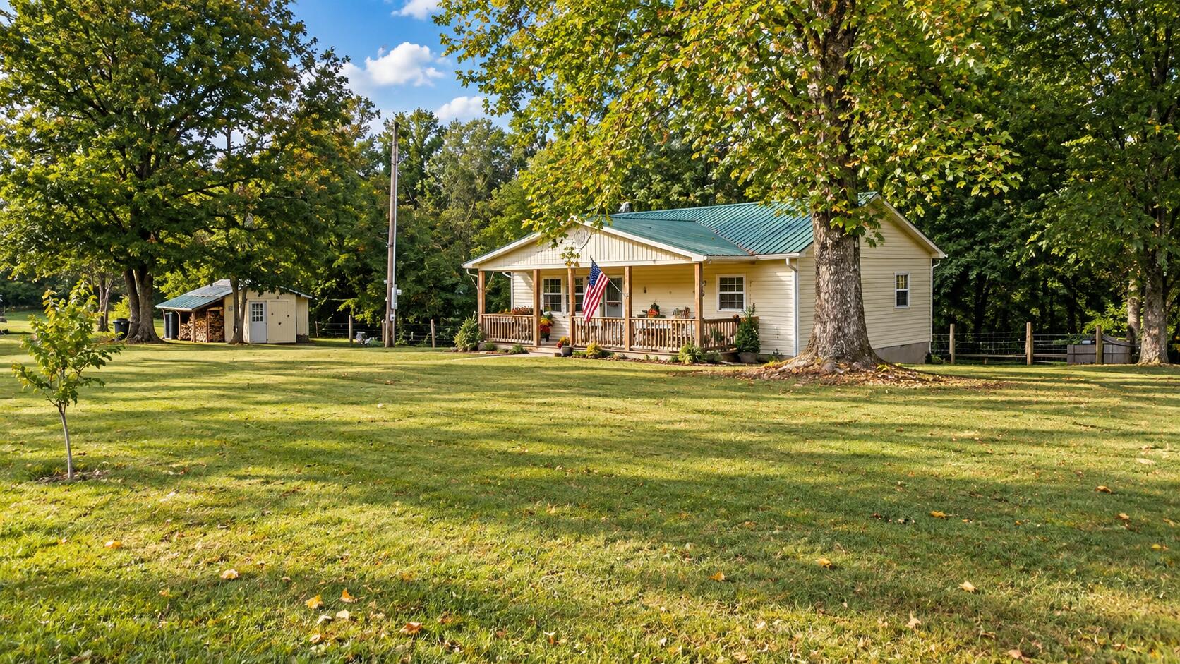 Cream-colored house with a green metal roof, covered porch, American flag, and detached shed among mature trees.