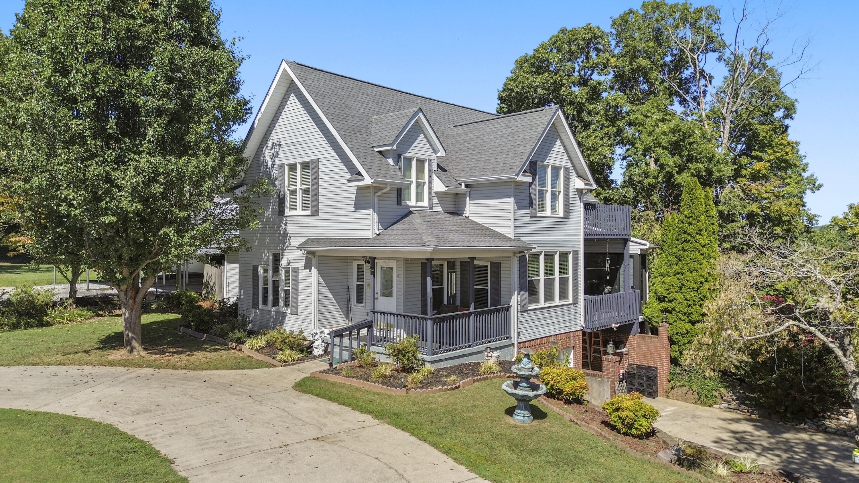 Gray two-story house with a covered porch, raised deck, curved driveway, and tiered lawn fountain.
