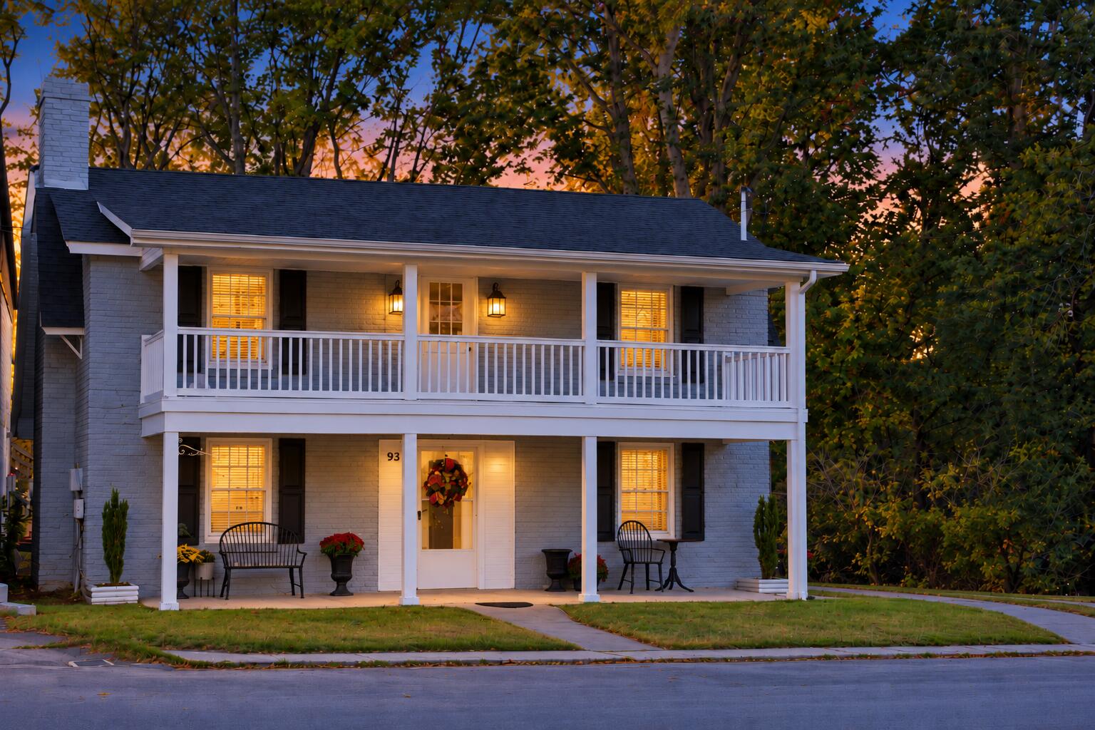 Gray brick two-story house with a white double-decker porch glowing at dusk beneath autumn trees.