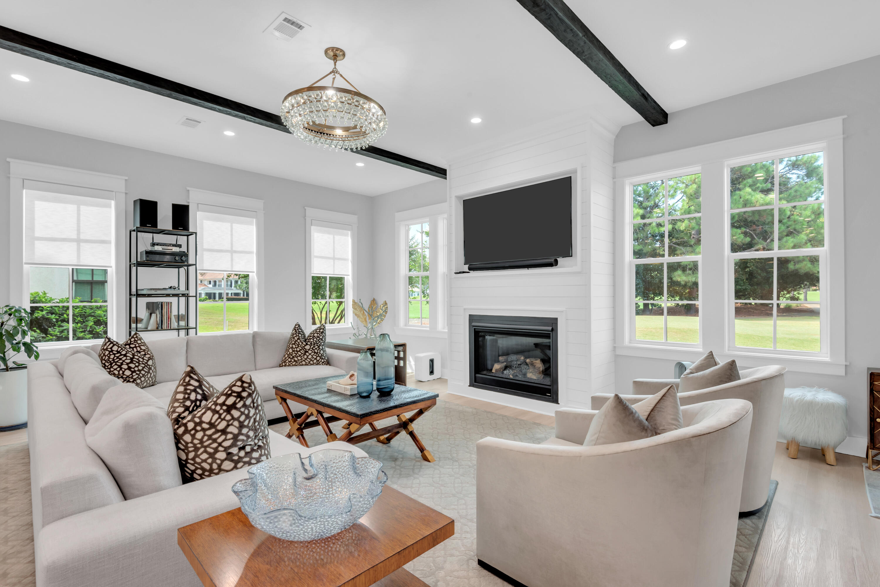 Living room with a white fireplace, mounted TV, dark ceiling beams, and large windows.