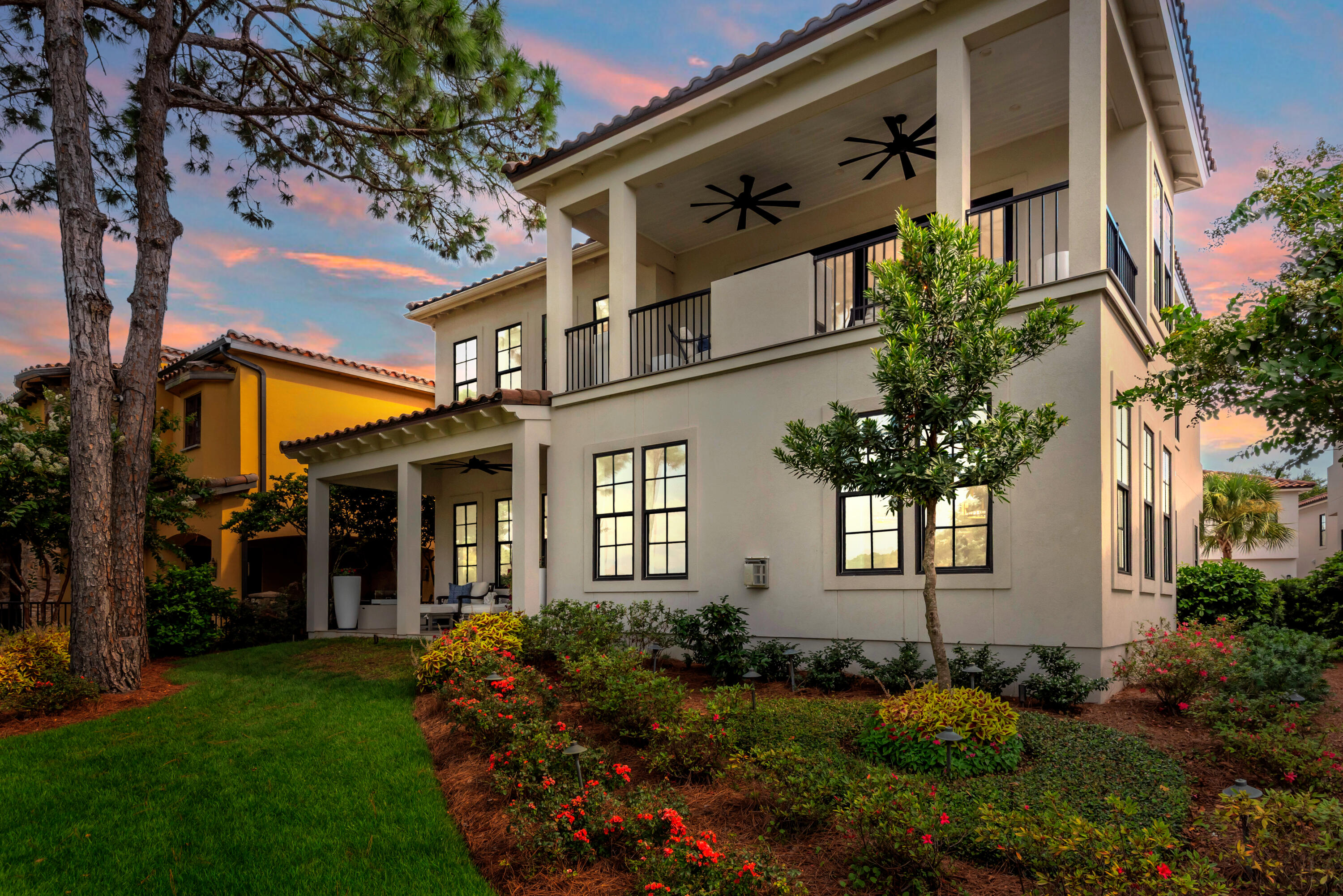 Two-story cream home with a covered patio and balcony, surrounded by landscaped gardens at sunset.