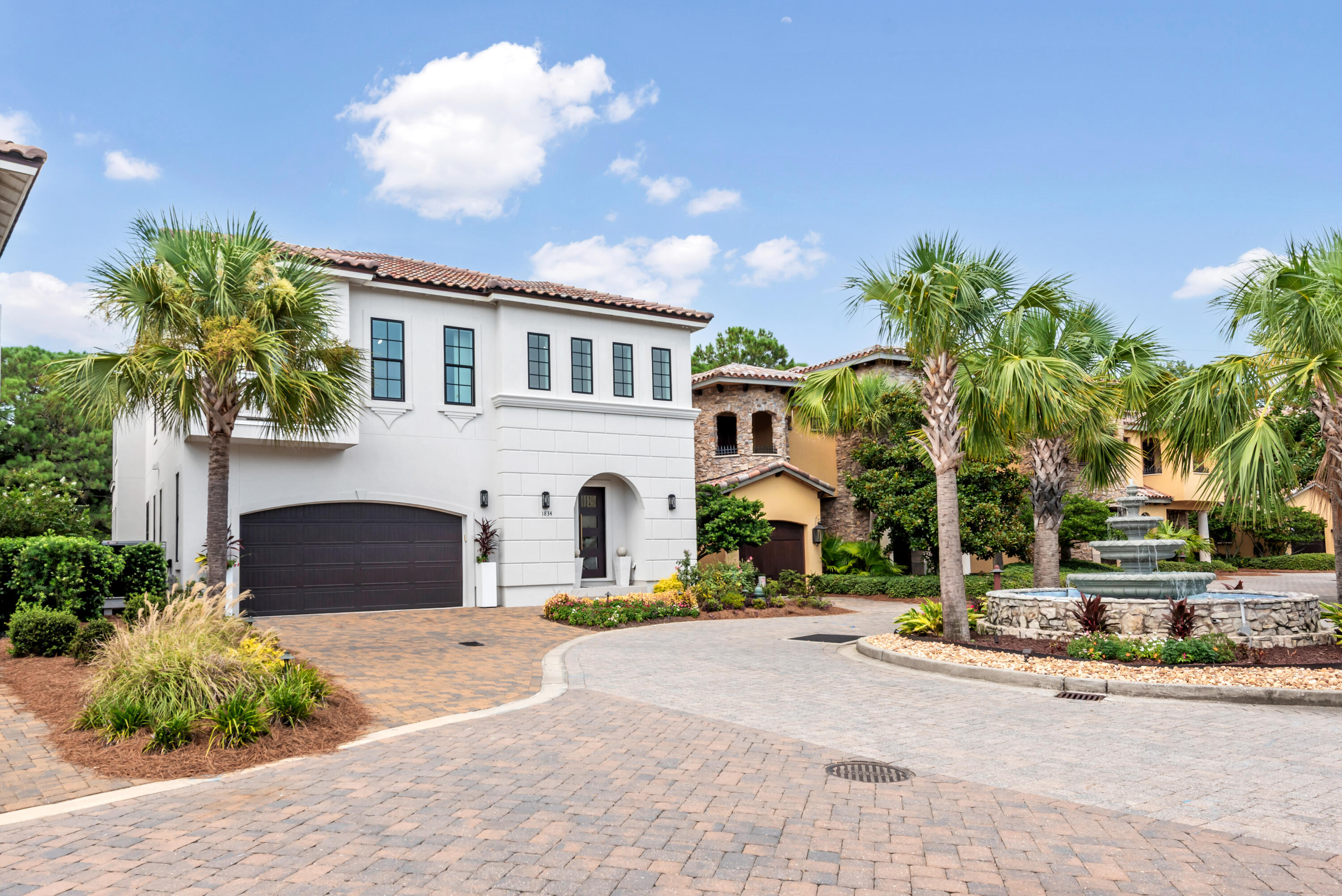 Two-story white home with a dark double garage, palm trees, and a courtyard fountain.