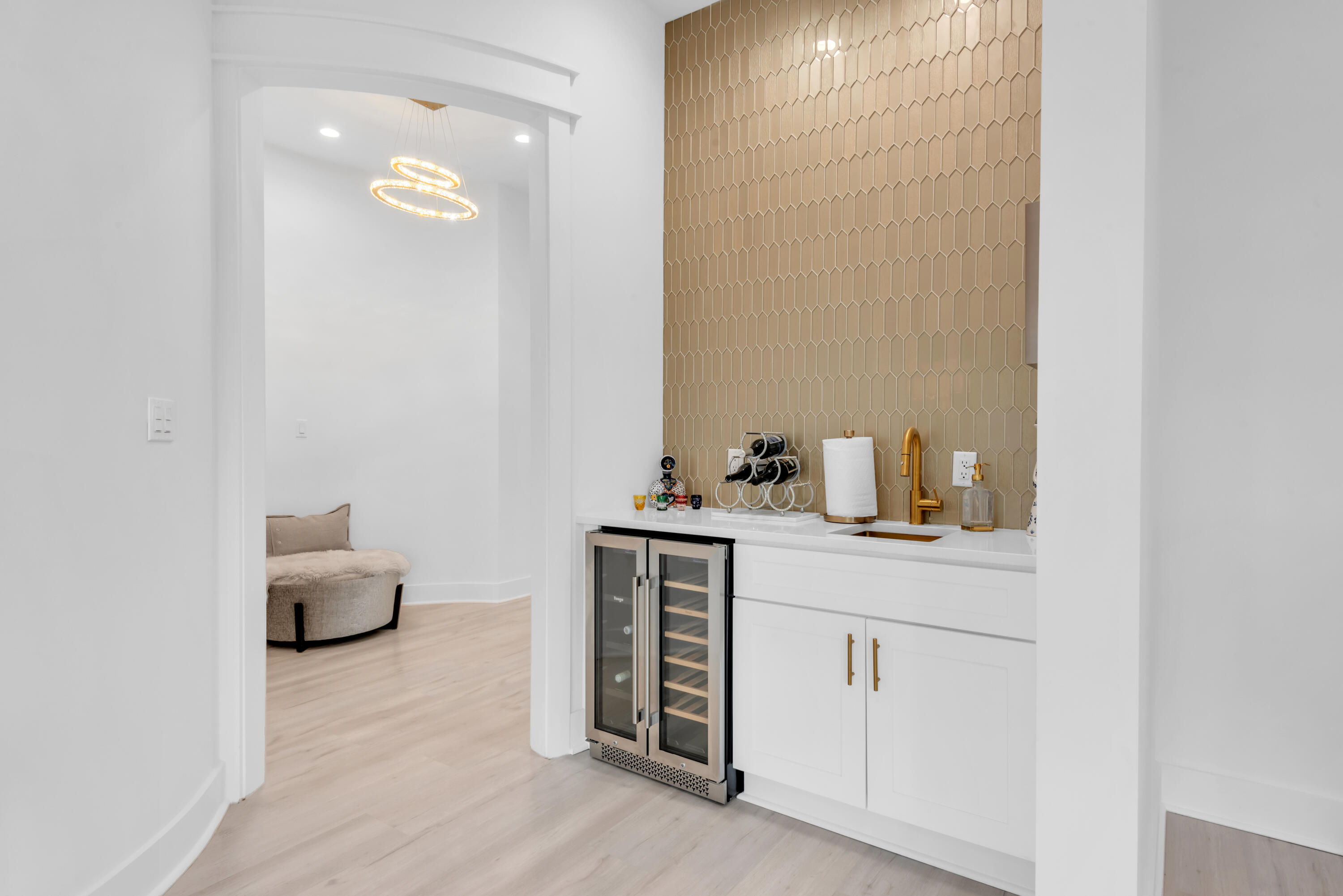 Home beverage bar with wine refrigerator and gold fixtures beside an arched opening to a sitting area.