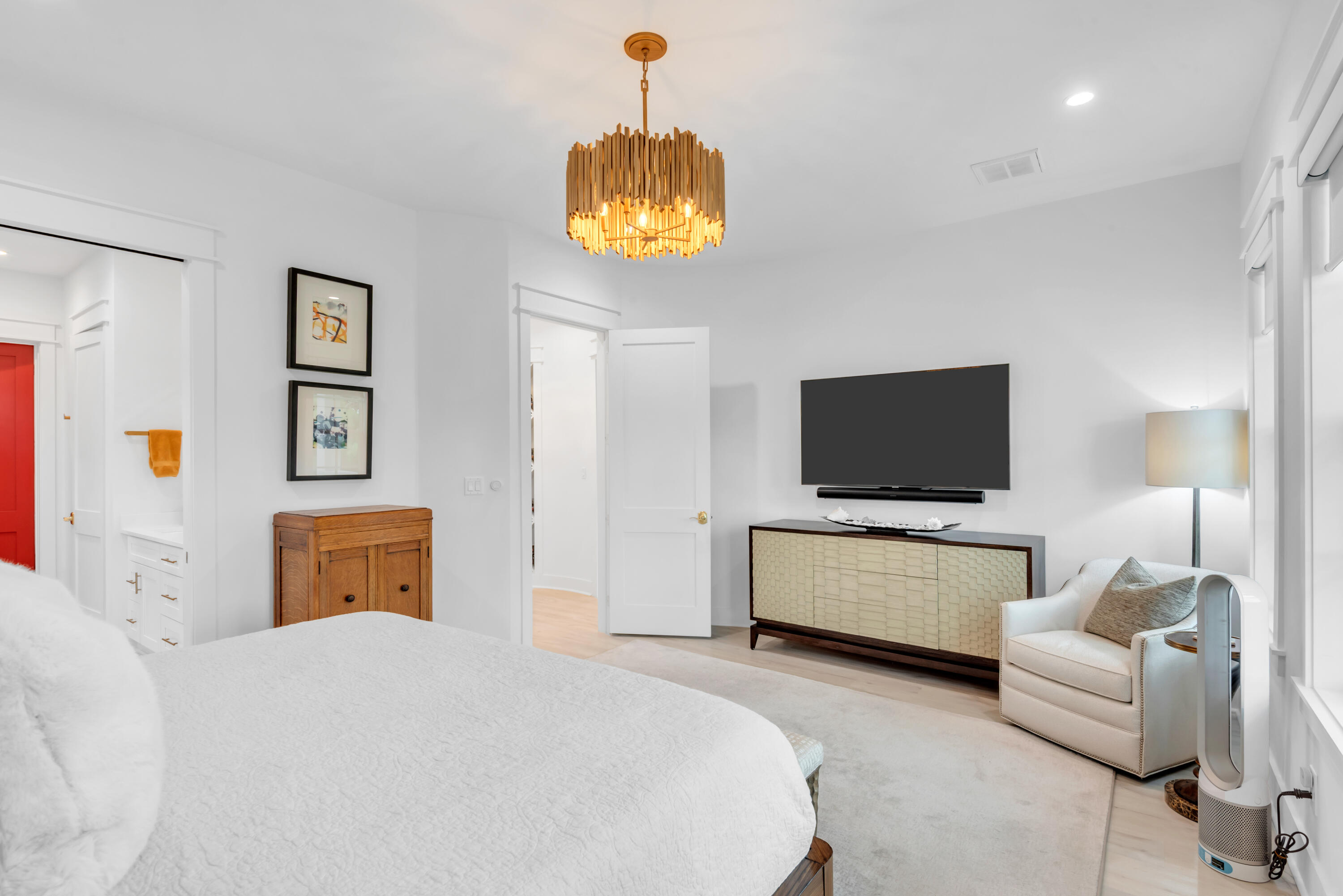 Bedroom with a large white bed, wall-mounted TV, wood chandelier, and armchair beside tall windows.