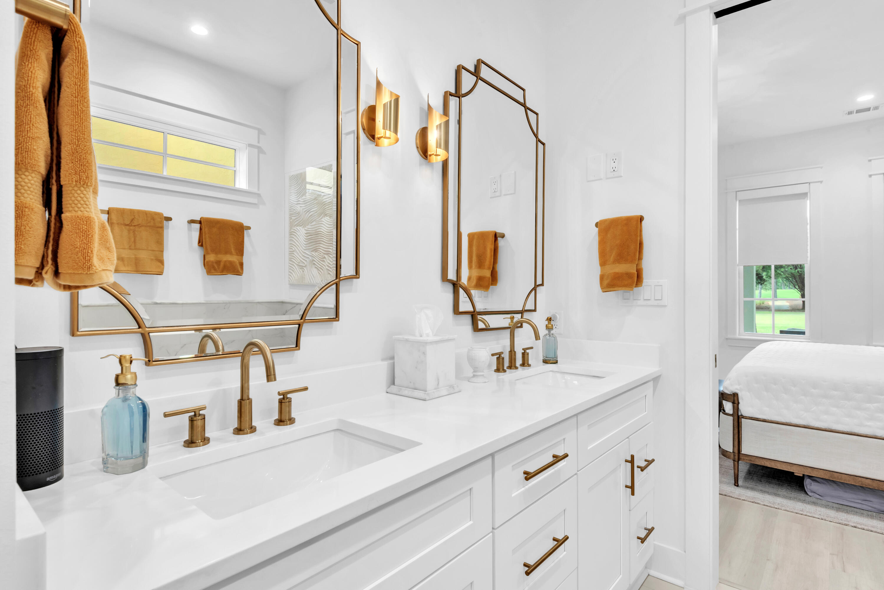 Bright double-vanity bathroom with gold fixtures and framed mirrors beside a bedroom doorway.