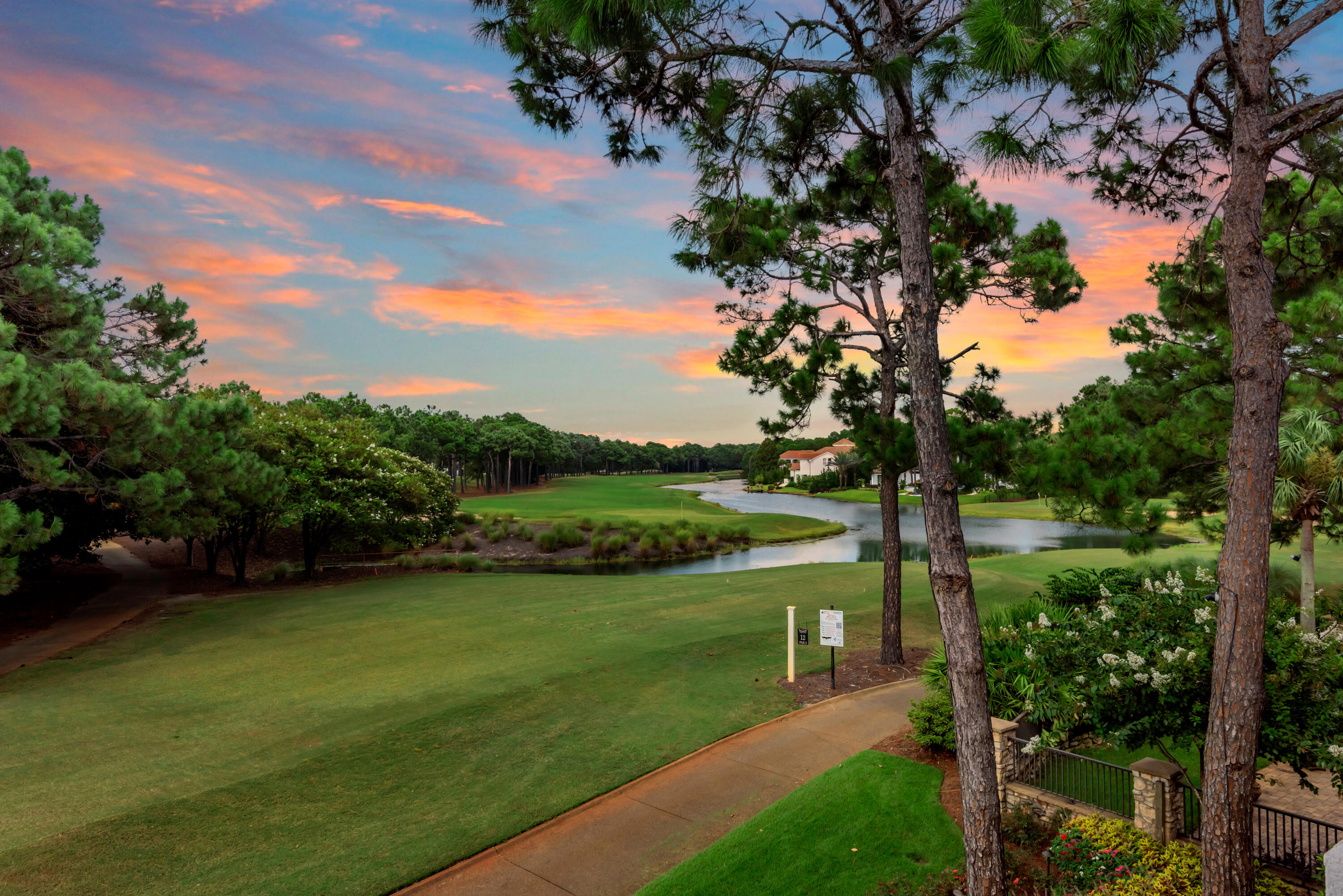 Golf course and winding pond framed by pine trees beneath a colorful sunset sky.