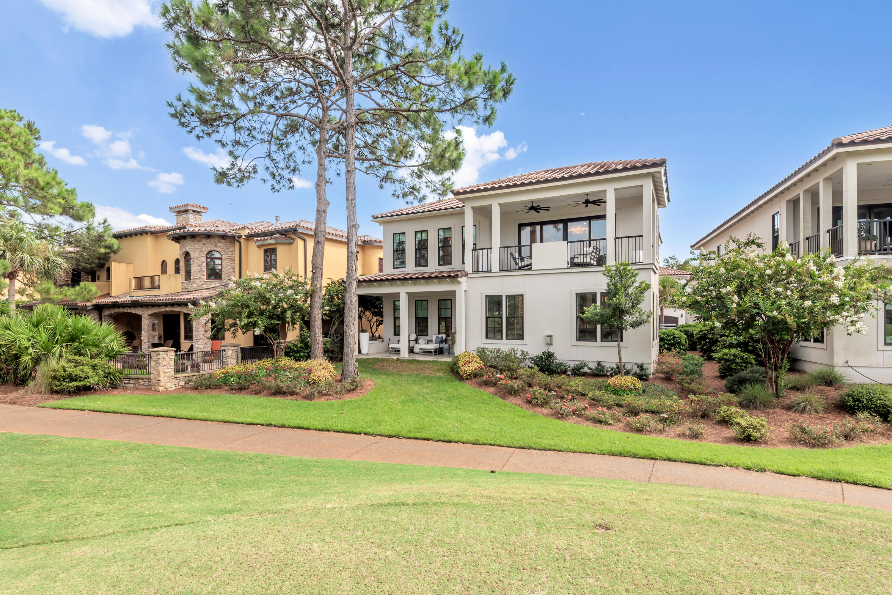 White two-story home with a covered balcony, tile roof, and landscaped lawn beneath tall pine trees.