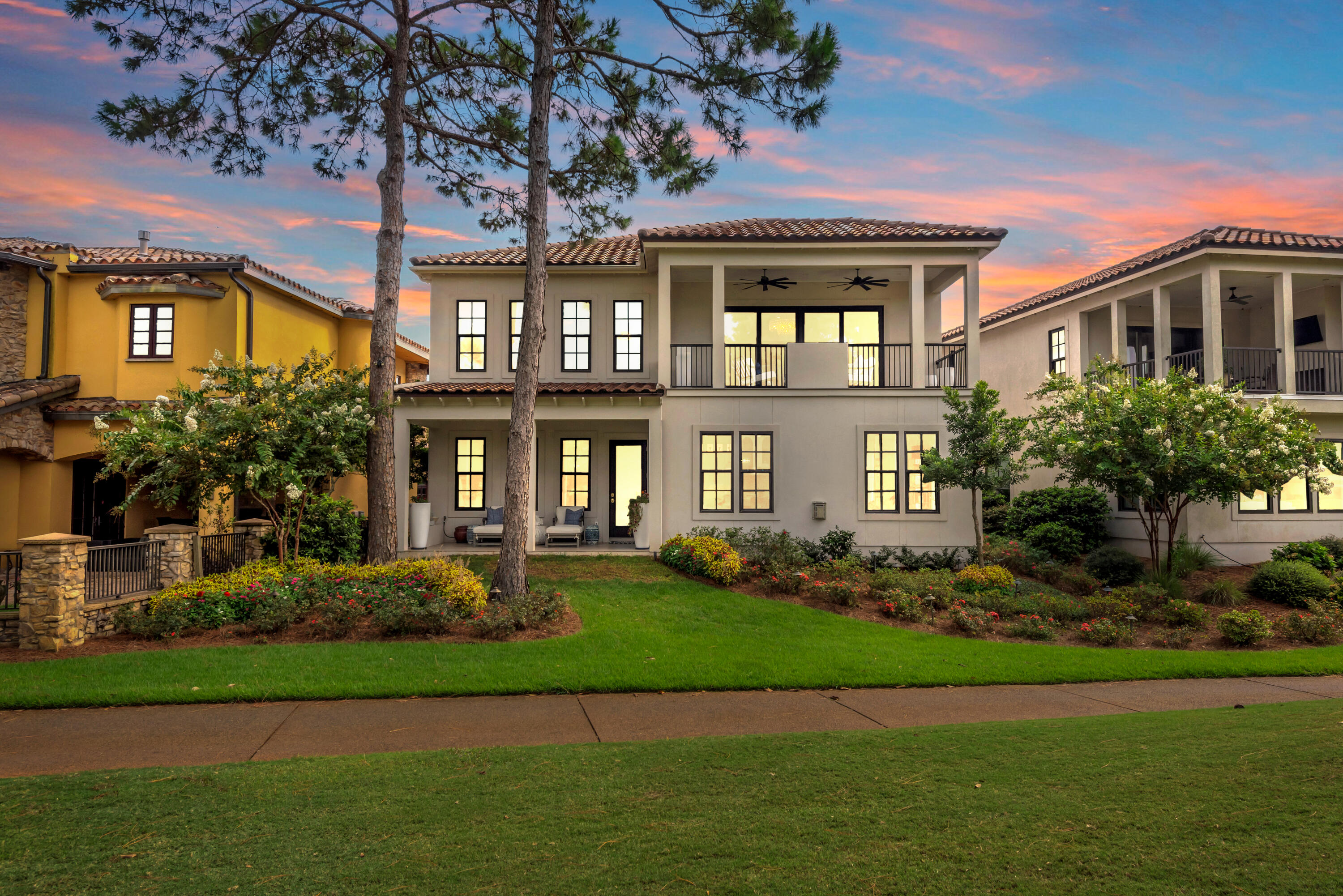 Two-story cream residence with a covered balcony, flowering landscaping, and neighboring homes at dusk.