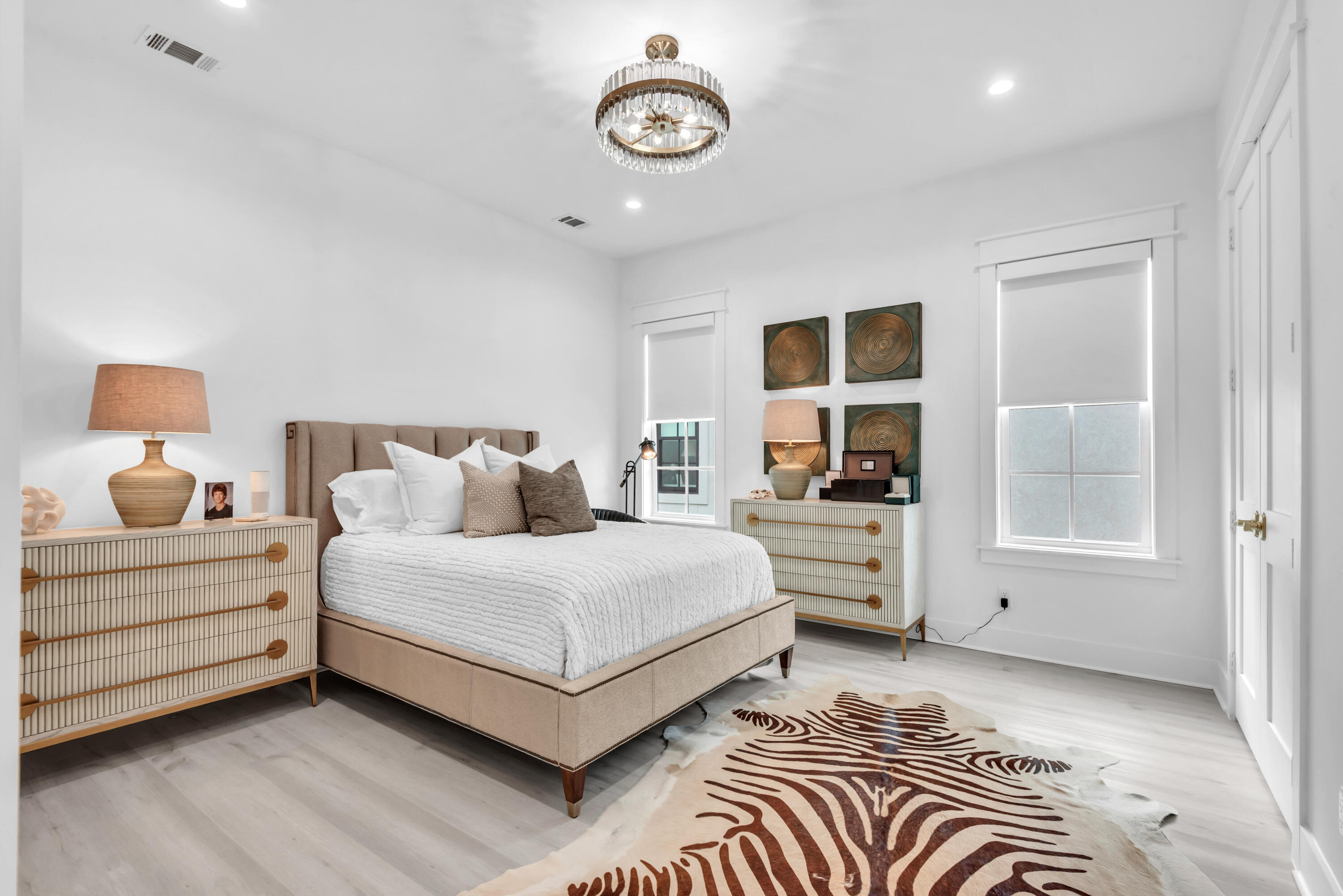 Staged bedroom with a beige bed, crystal chandelier, matching dressers, and patterned rug.