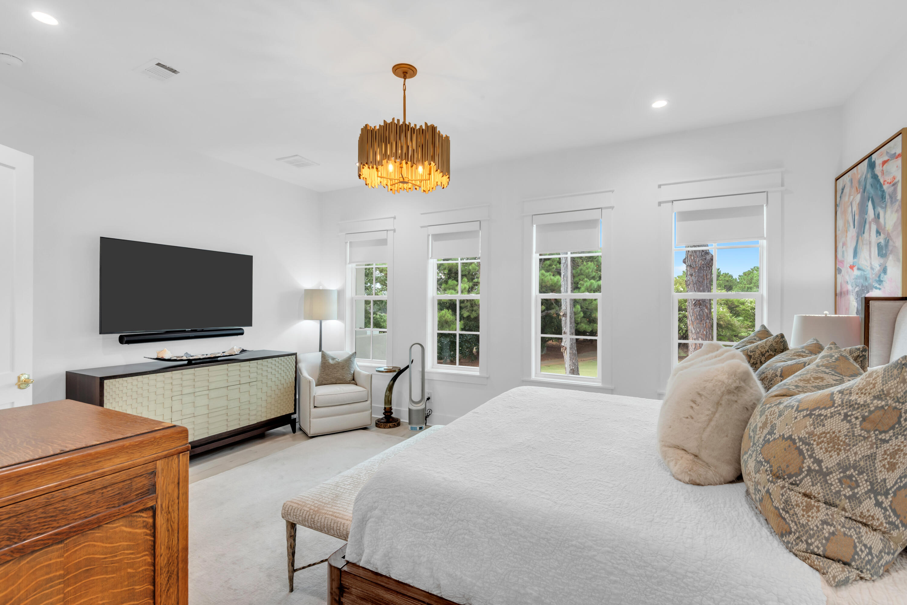 Bedroom with a large bed, patterned pillows, wall-mounted television, and windows overlooking trees.