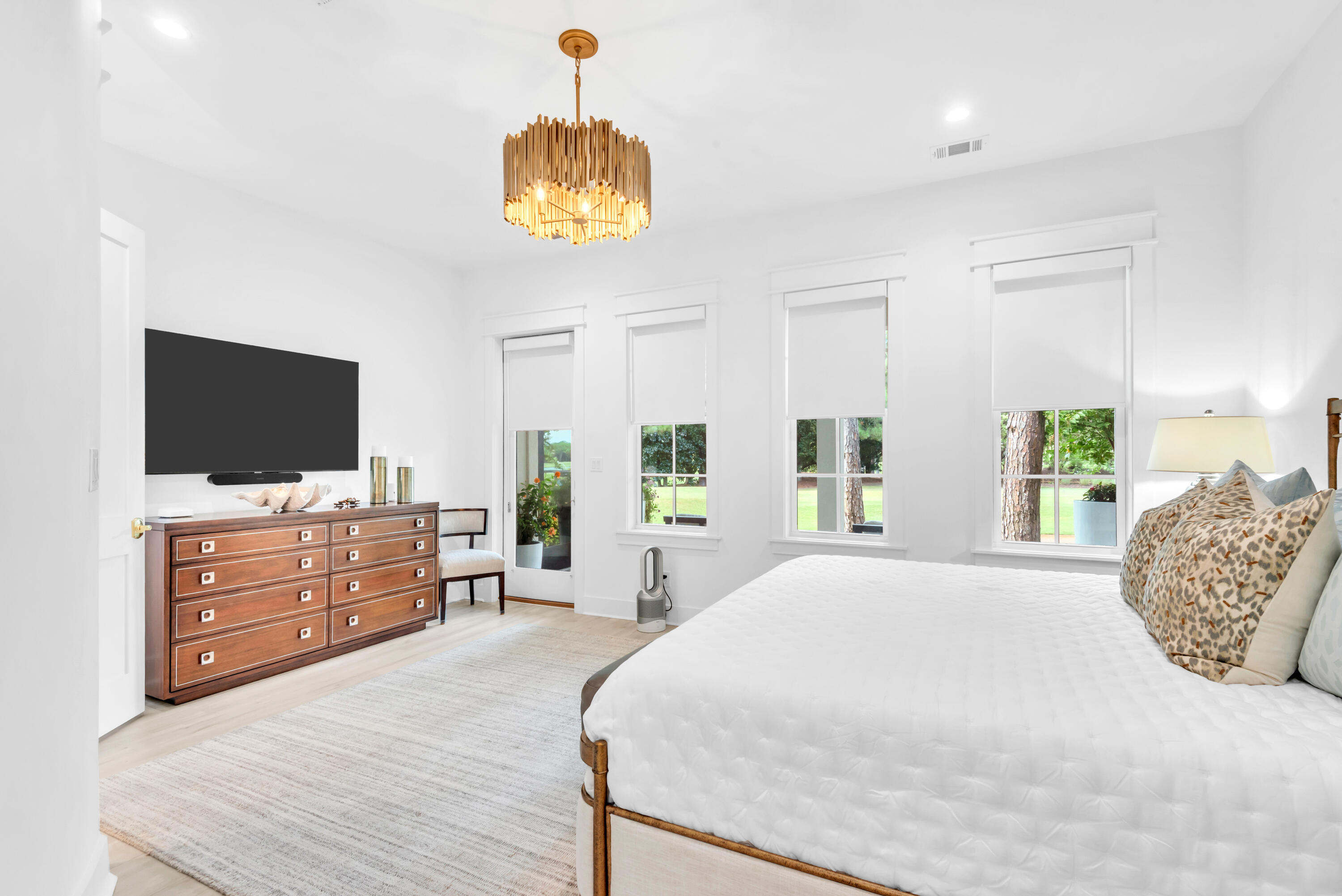 Bright bedroom with a white bed, wood dresser, wall-mounted television, and windows overlooking a green yard.