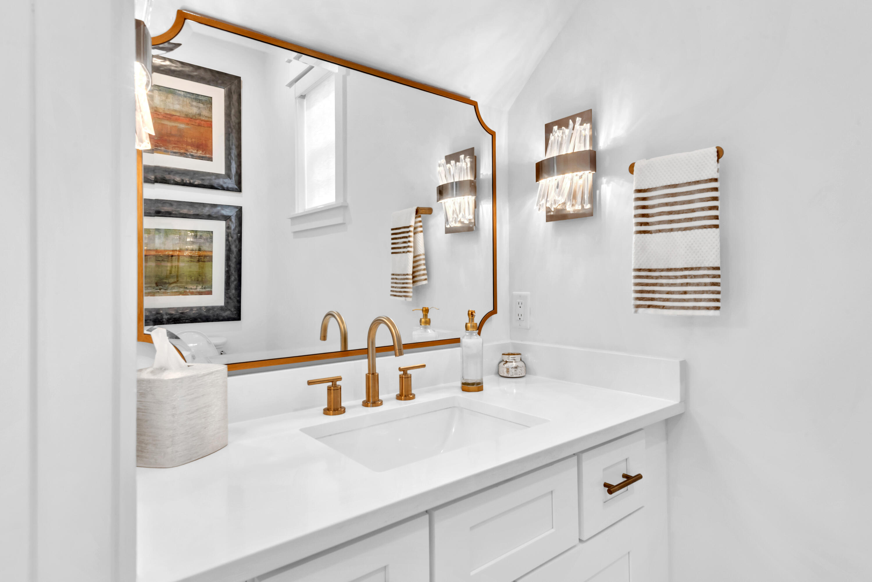 Bathroom vanity with a white sink, brass fixtures, gold-trimmed mirror, and crystal sconces.