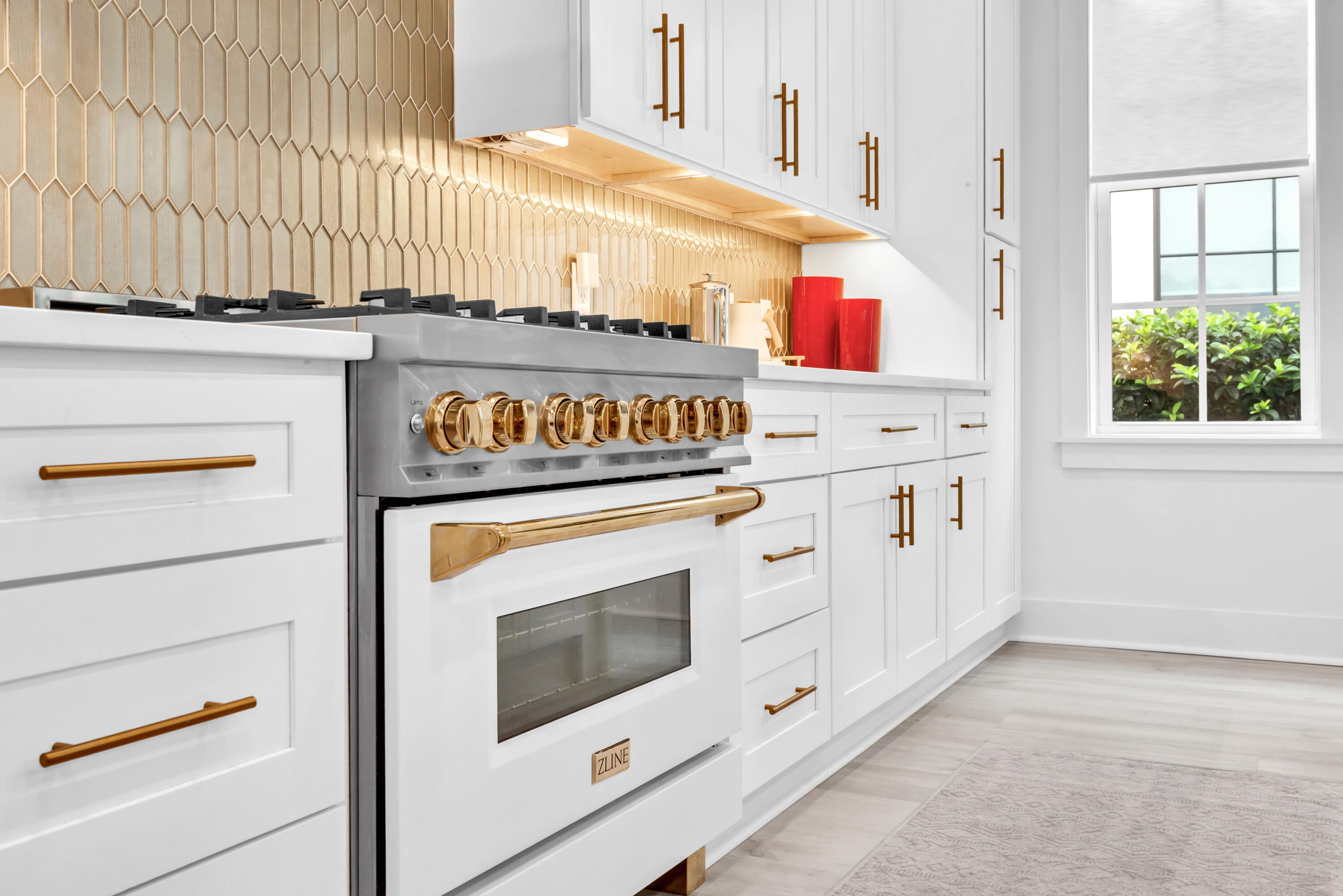 White kitchen with a brass-trimmed range, geometric tile backsplash, and window overlooking greenery.