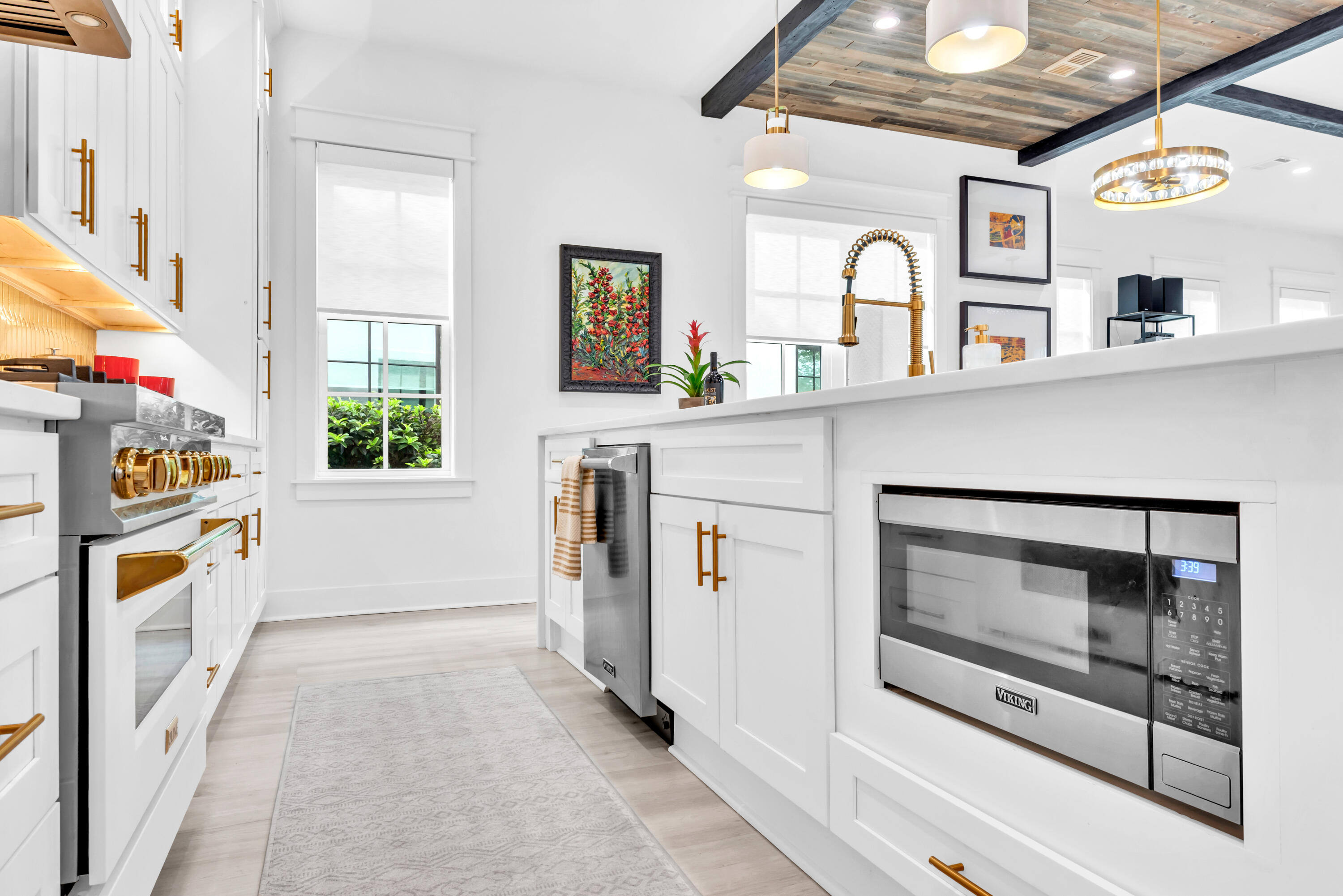 Bright white kitchen with gold fixtures, built-in Viking oven, large island, and wood-plank ceiling with dark beams.