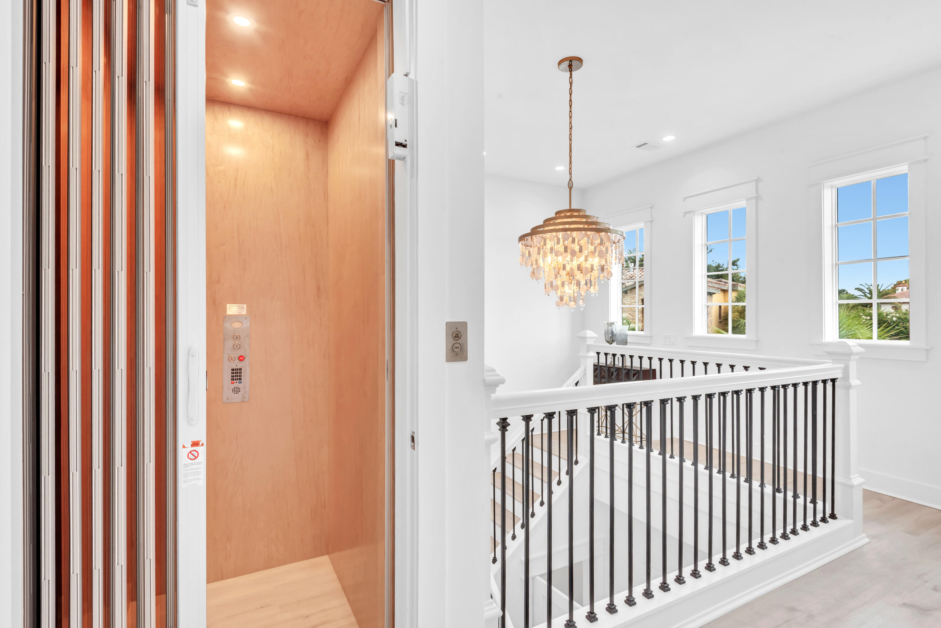Open wood-paneled elevator beside a staircase with dark iron balusters and a crystal chandelier.
