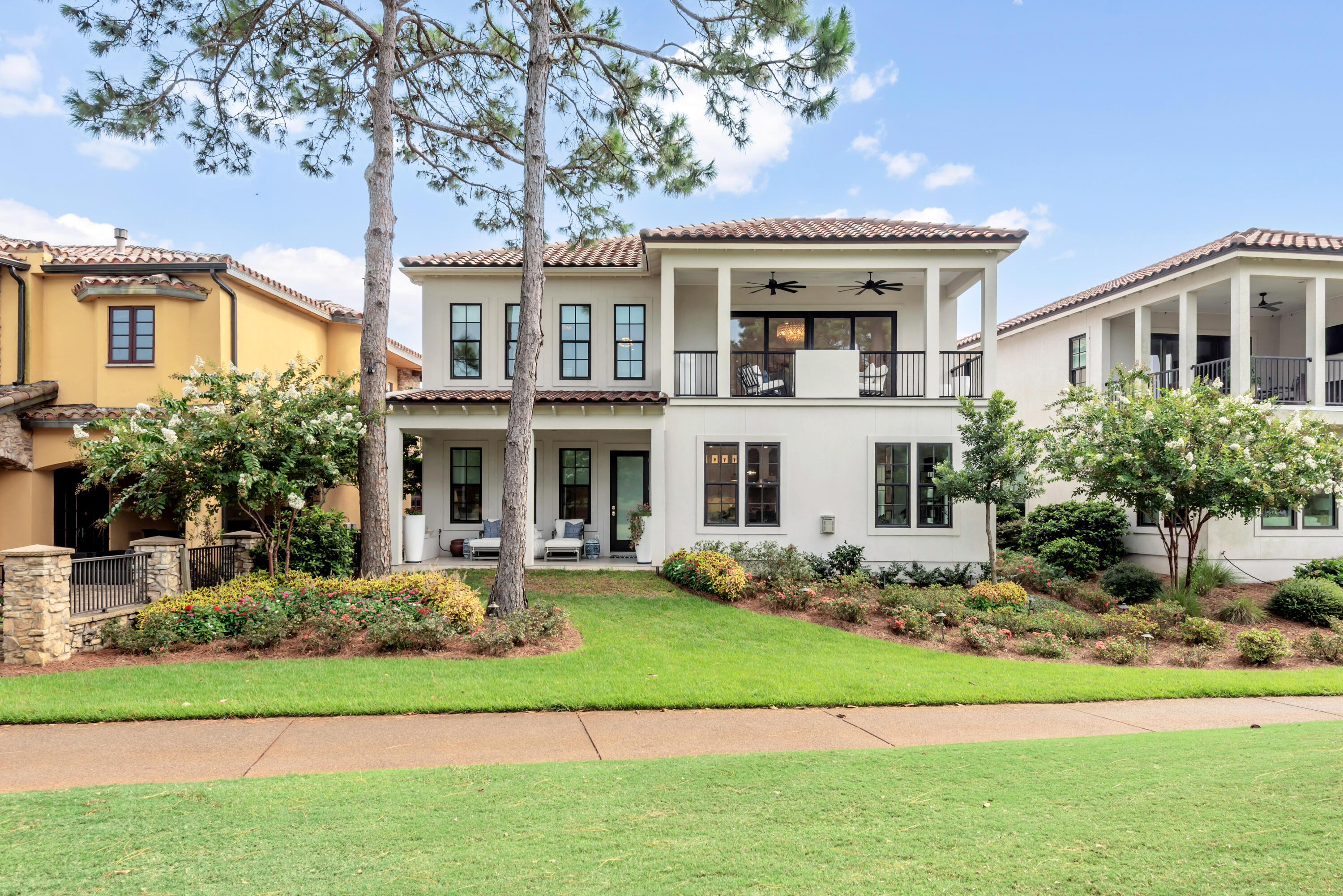 Two-story white stucco home with a covered balcony, landscaped garden, and tall pine trees.