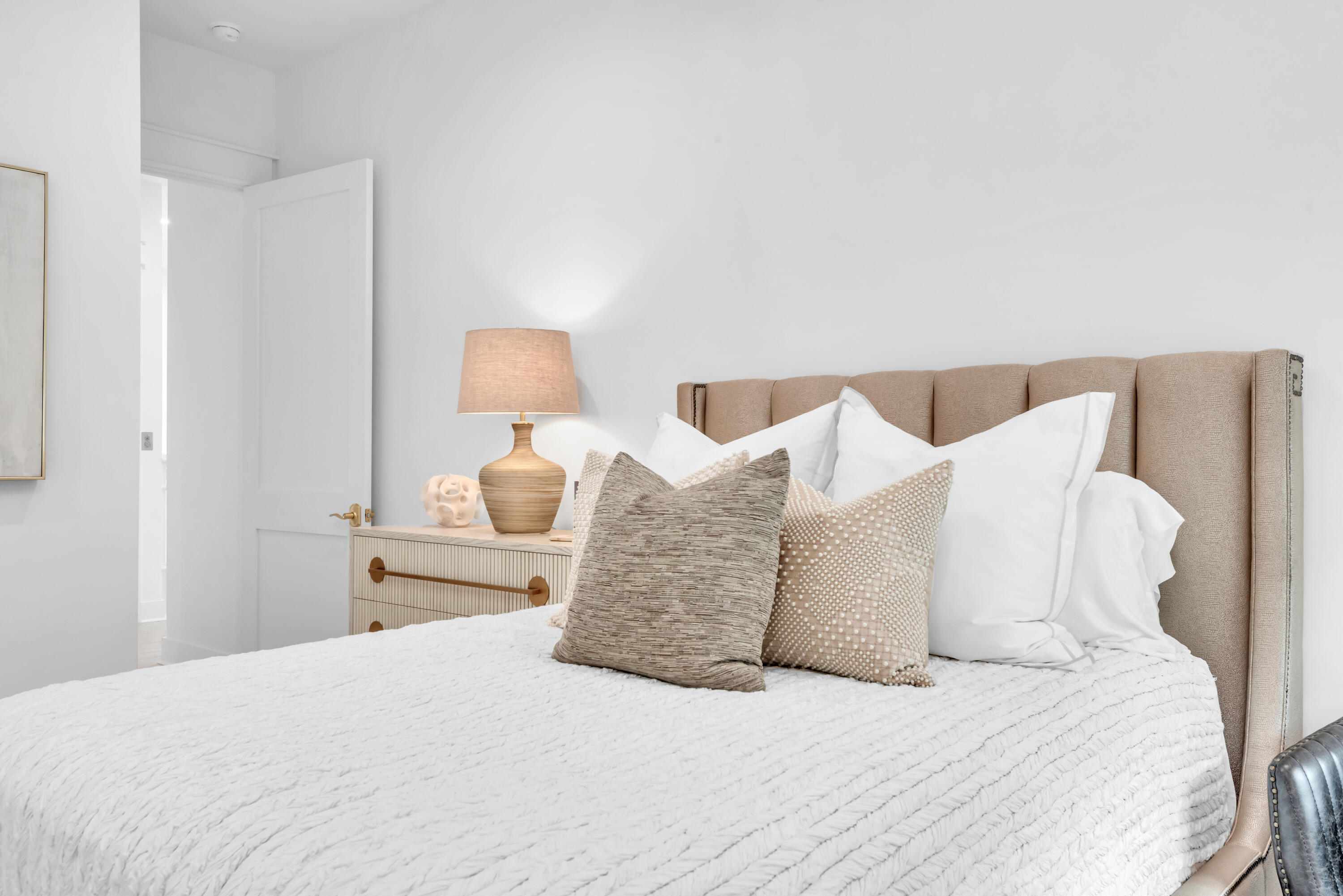 Bedroom with a beige upholstered headboard, layered neutral pillows, and a bedside lamp.