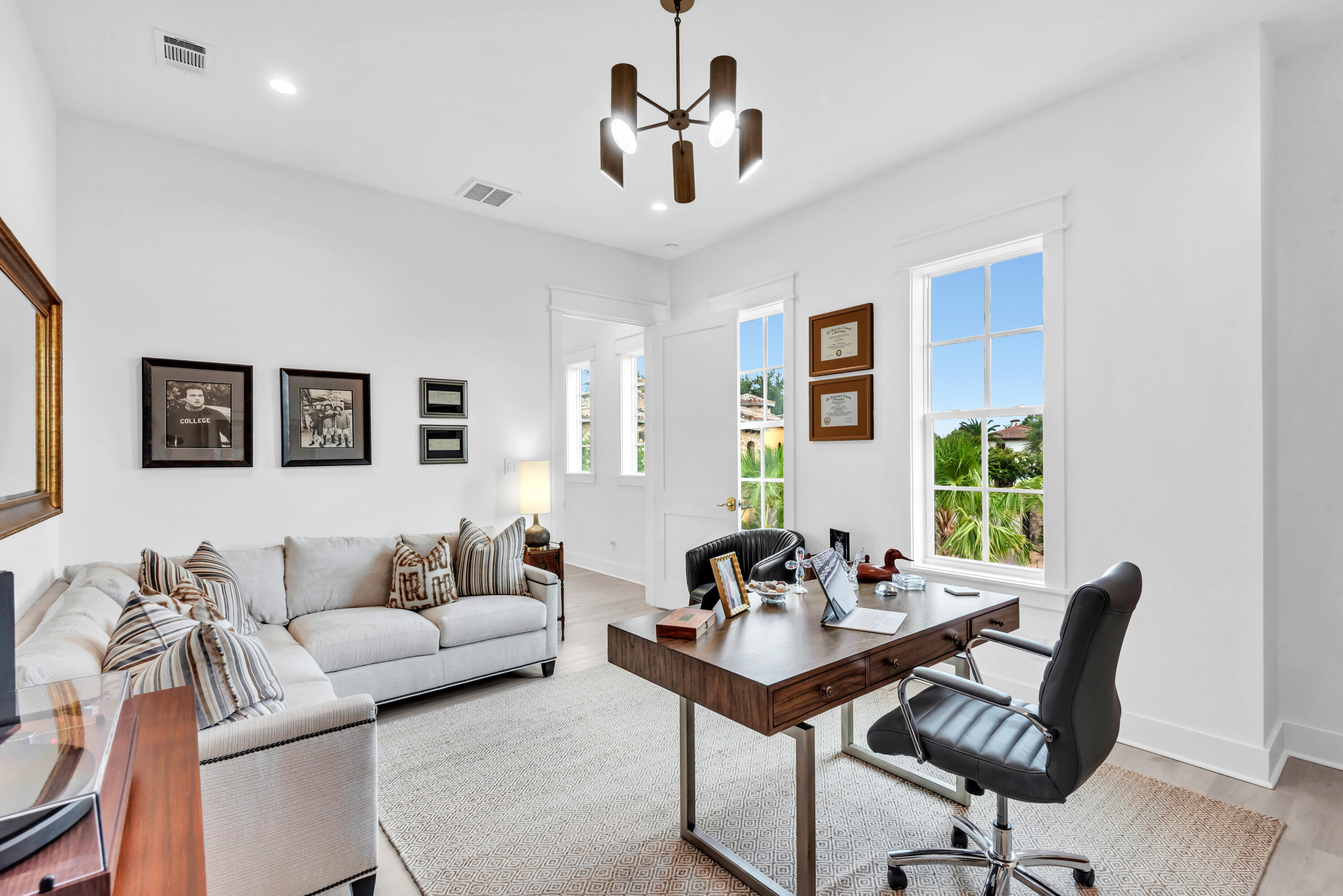 Bright home office with a dark wood desk, black swivel chair, light sectional, and windows overlooking tropical greenery.