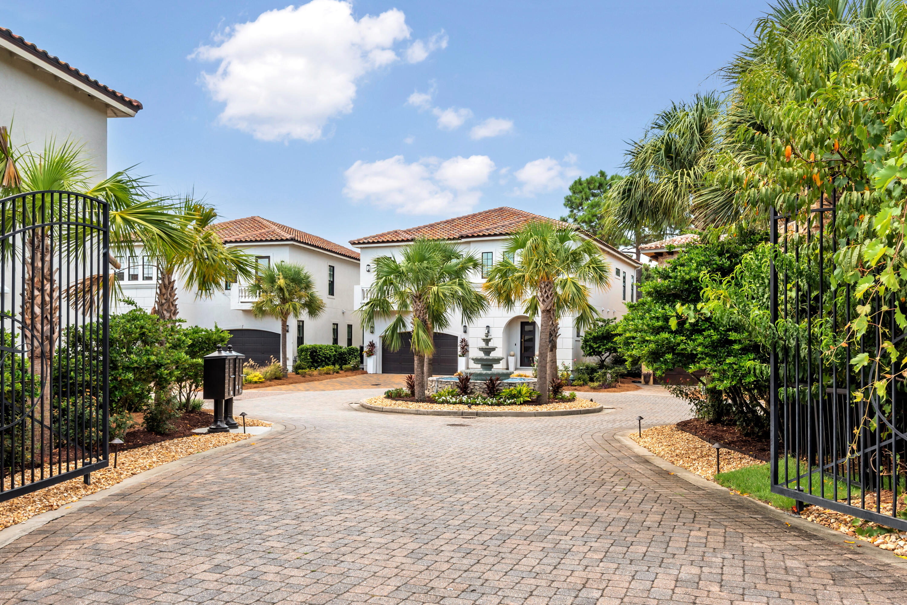 Gated courtyard with white stucco homes, palm trees, and a fountain along a brick-paved drive.