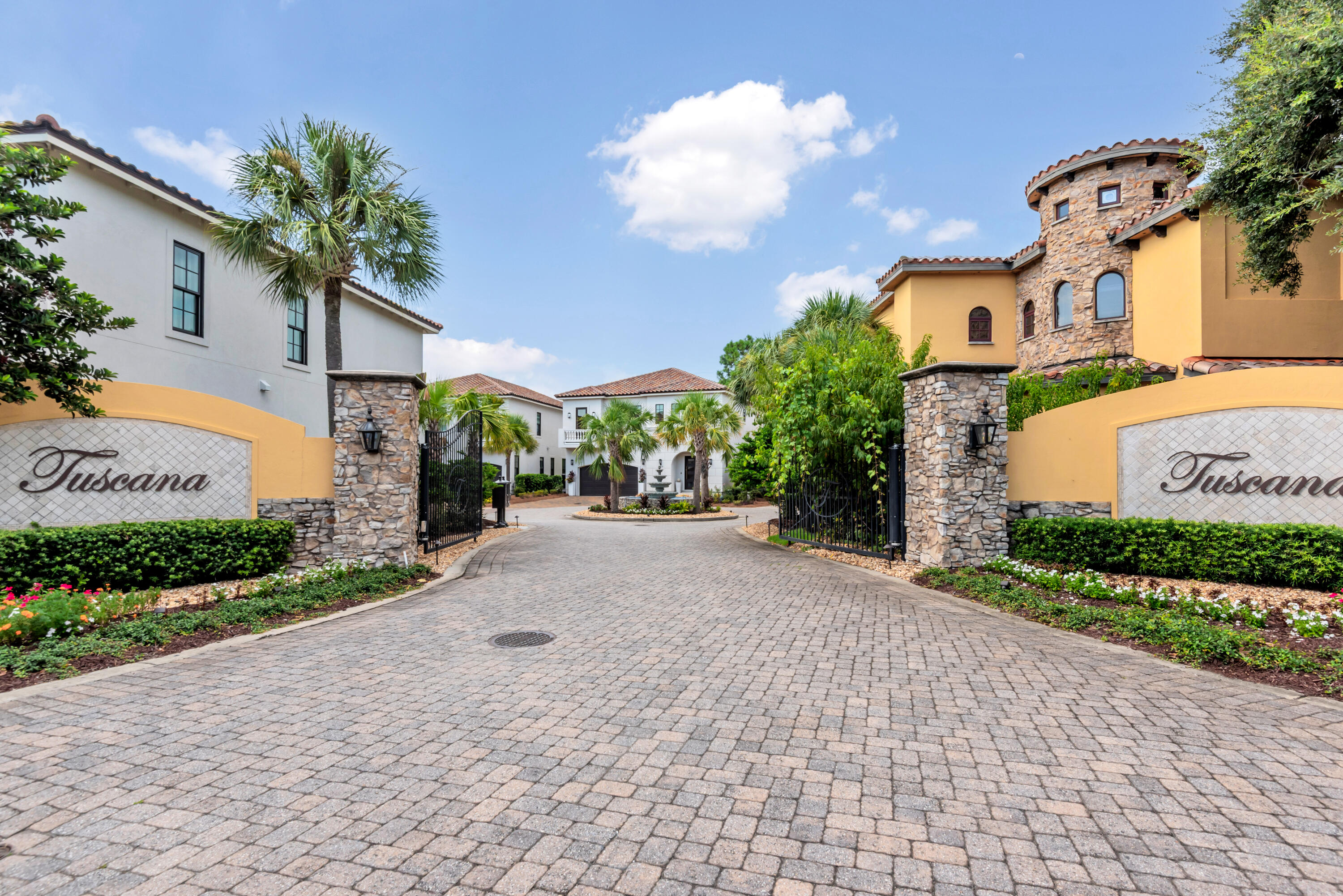 Gated Tuscana entrance with stone pillars, open iron gates, palm trees, and a cobblestone driveway.