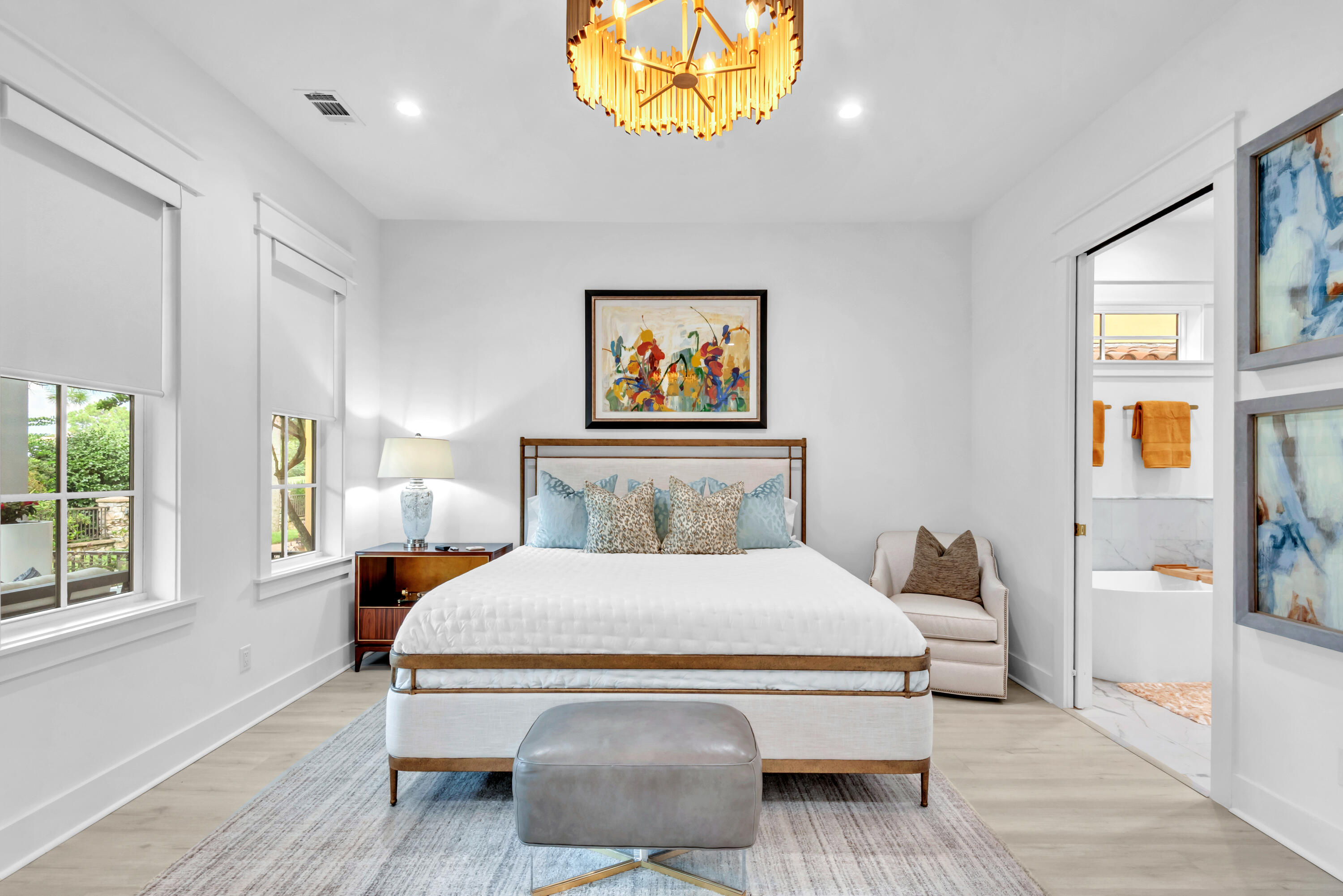 Staged bedroom with a white bed, gold chandelier, abstract artwork, and ensuite bathroom doorway.