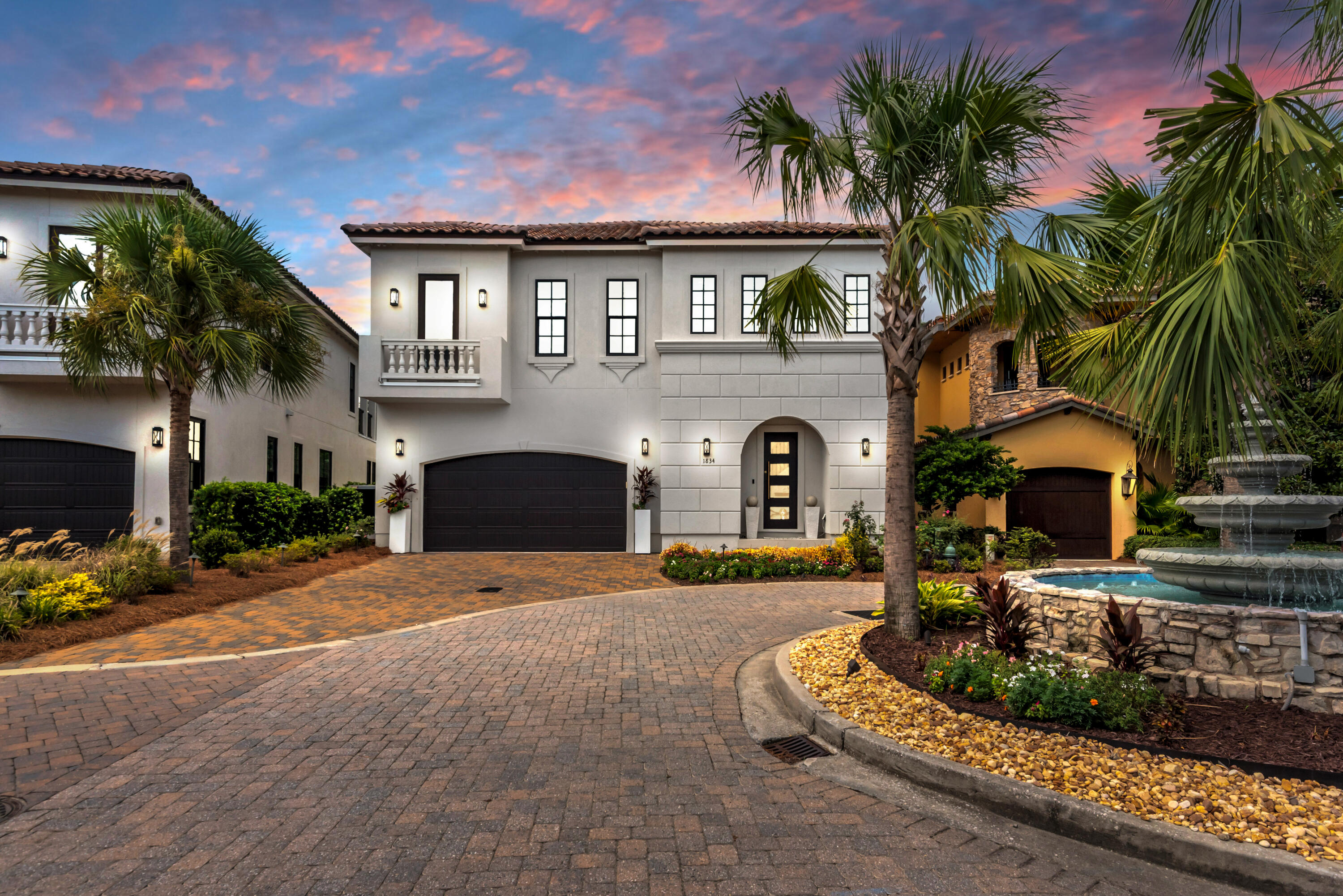 Two-story white home with a brick driveway, palm trees, and stone fountain at dusk.
