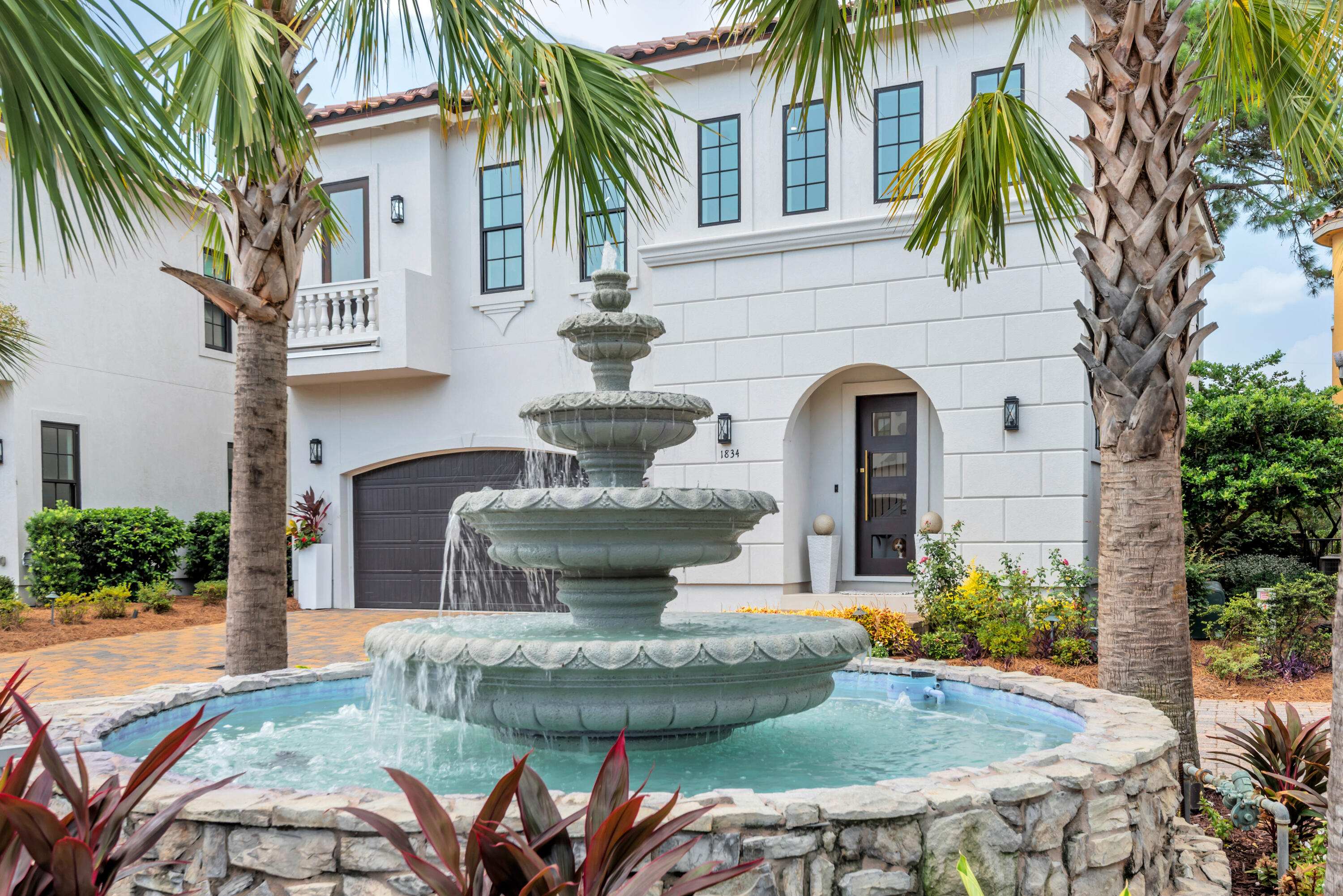 Two-story white stucco home with a tiered fountain, palm trees, and arched entry.