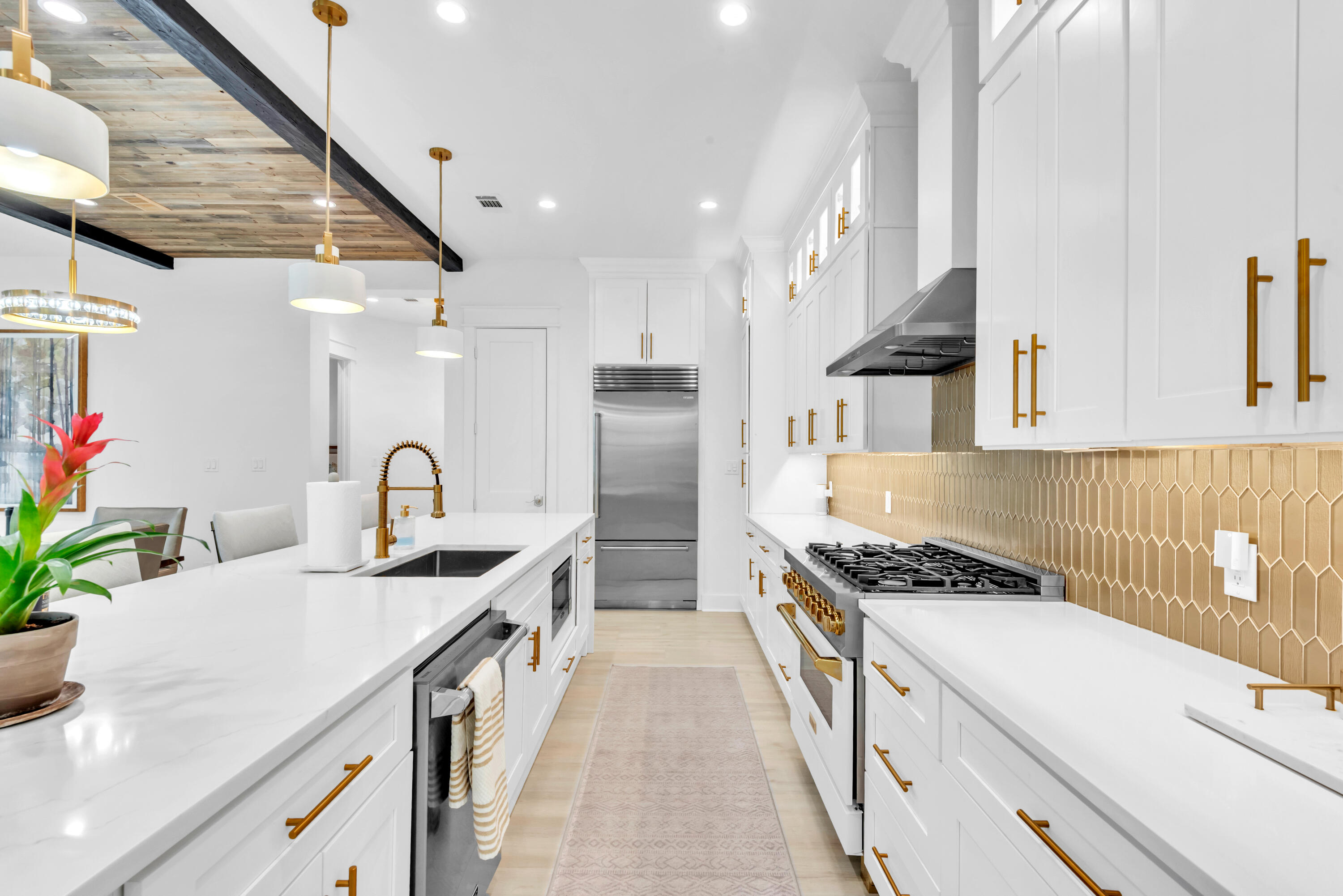 Bright white kitchen with a quartz island, gold tile backsplash, brass fixtures, and rustic wood ceiling detail.