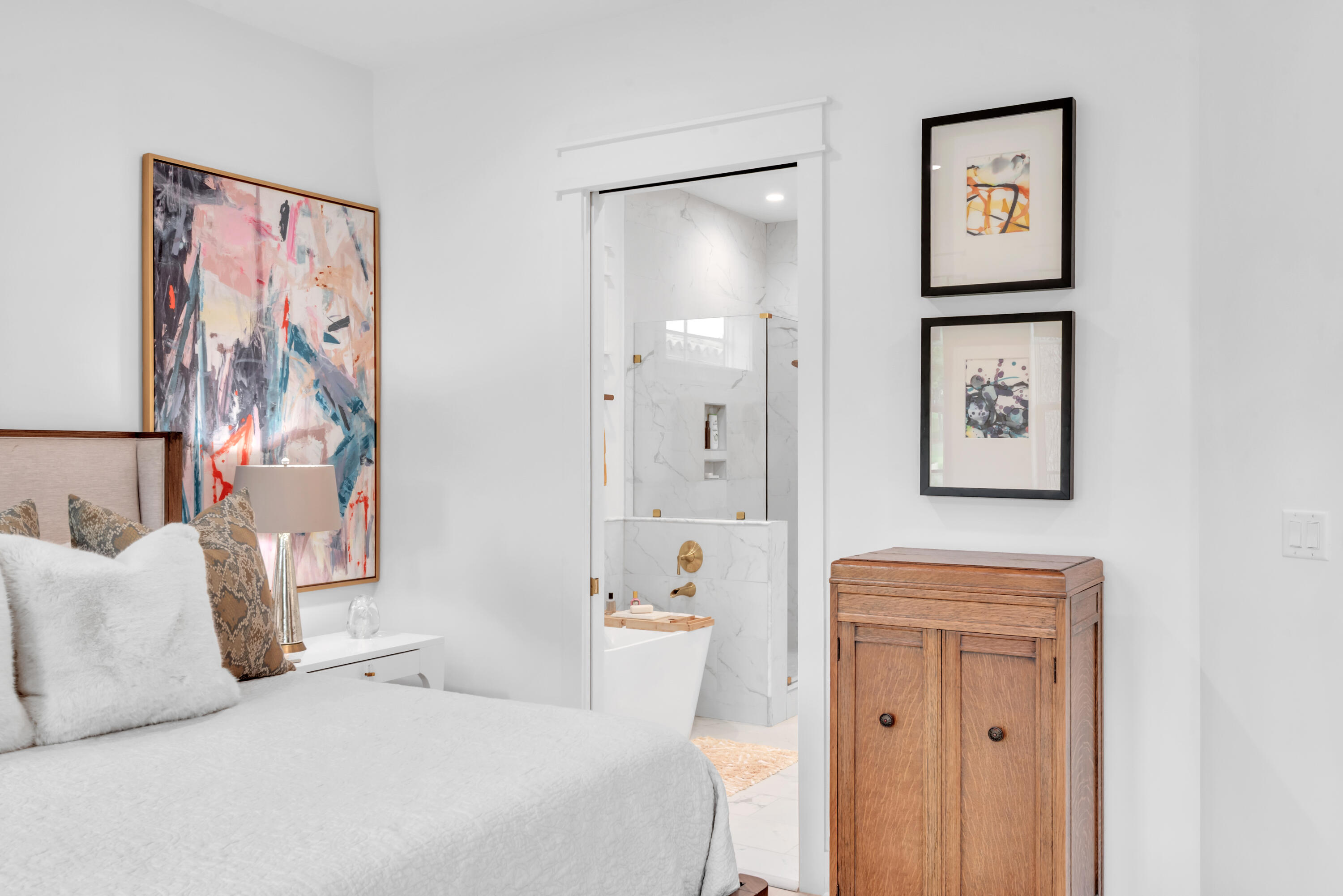 Bedroom with a white bed and colorful artwork beside an open marble-tiled shower.