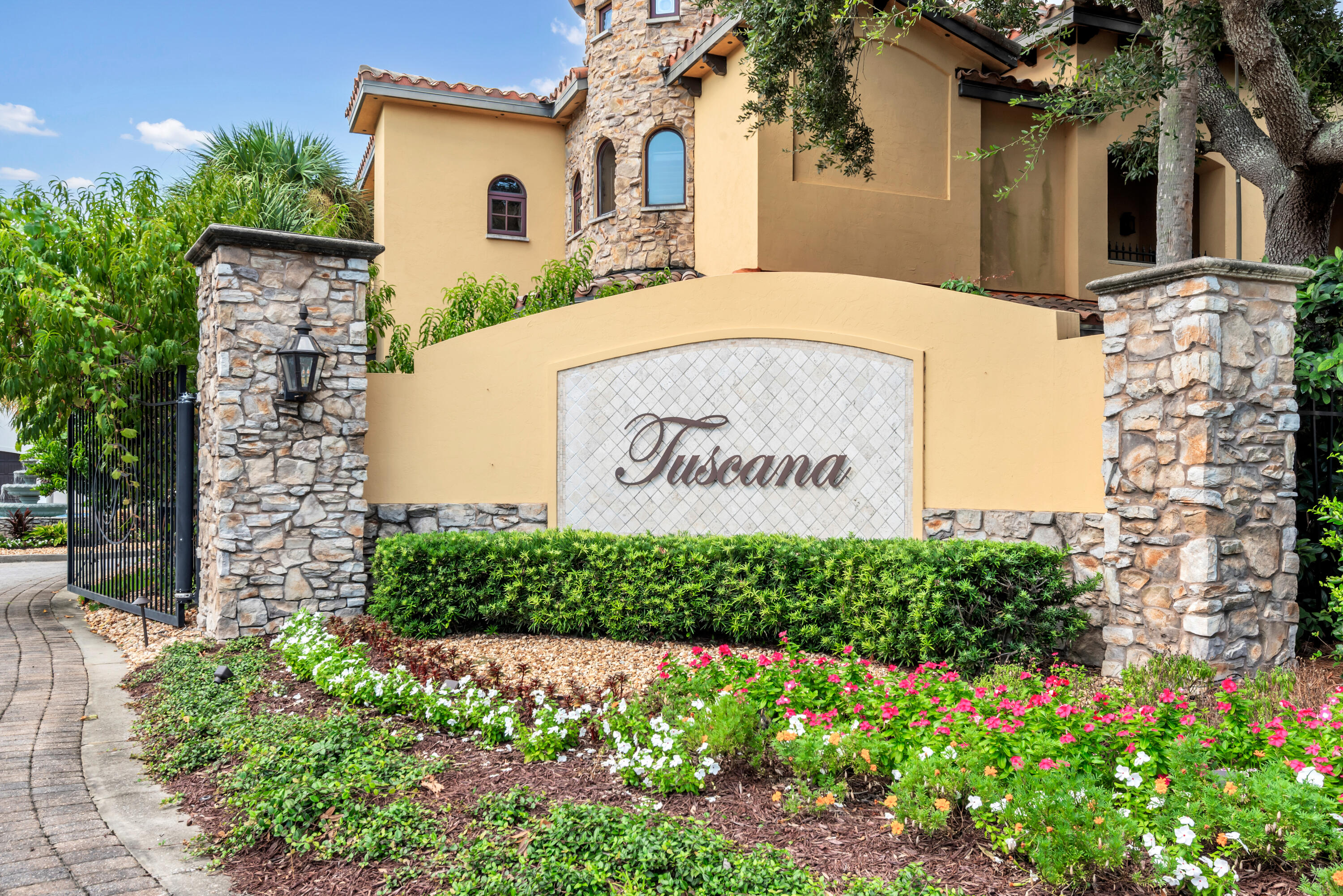 Tuscana community entrance sign beside a stone-and-stucco building and landscaped flower beds.