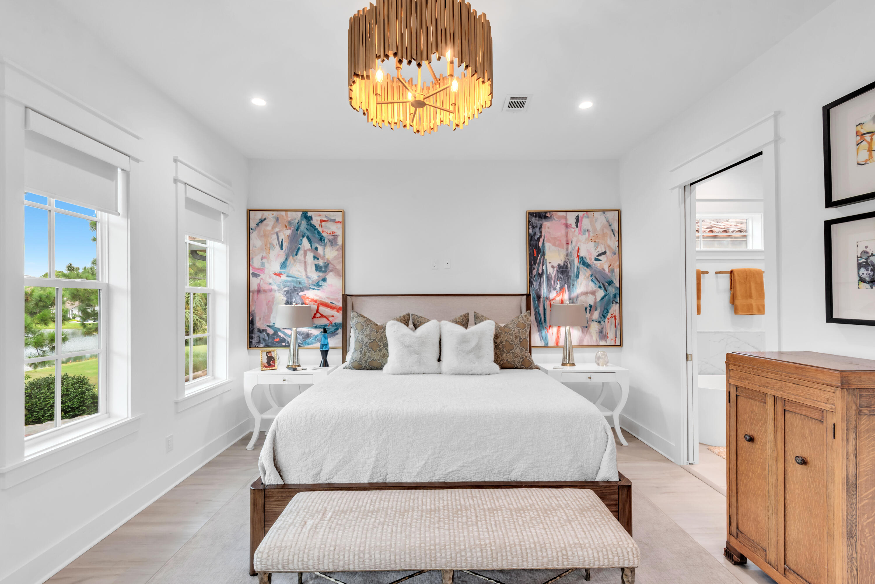 Staged bedroom with a white bed, colorful abstract art, wood chandelier, and windows overlooking water.