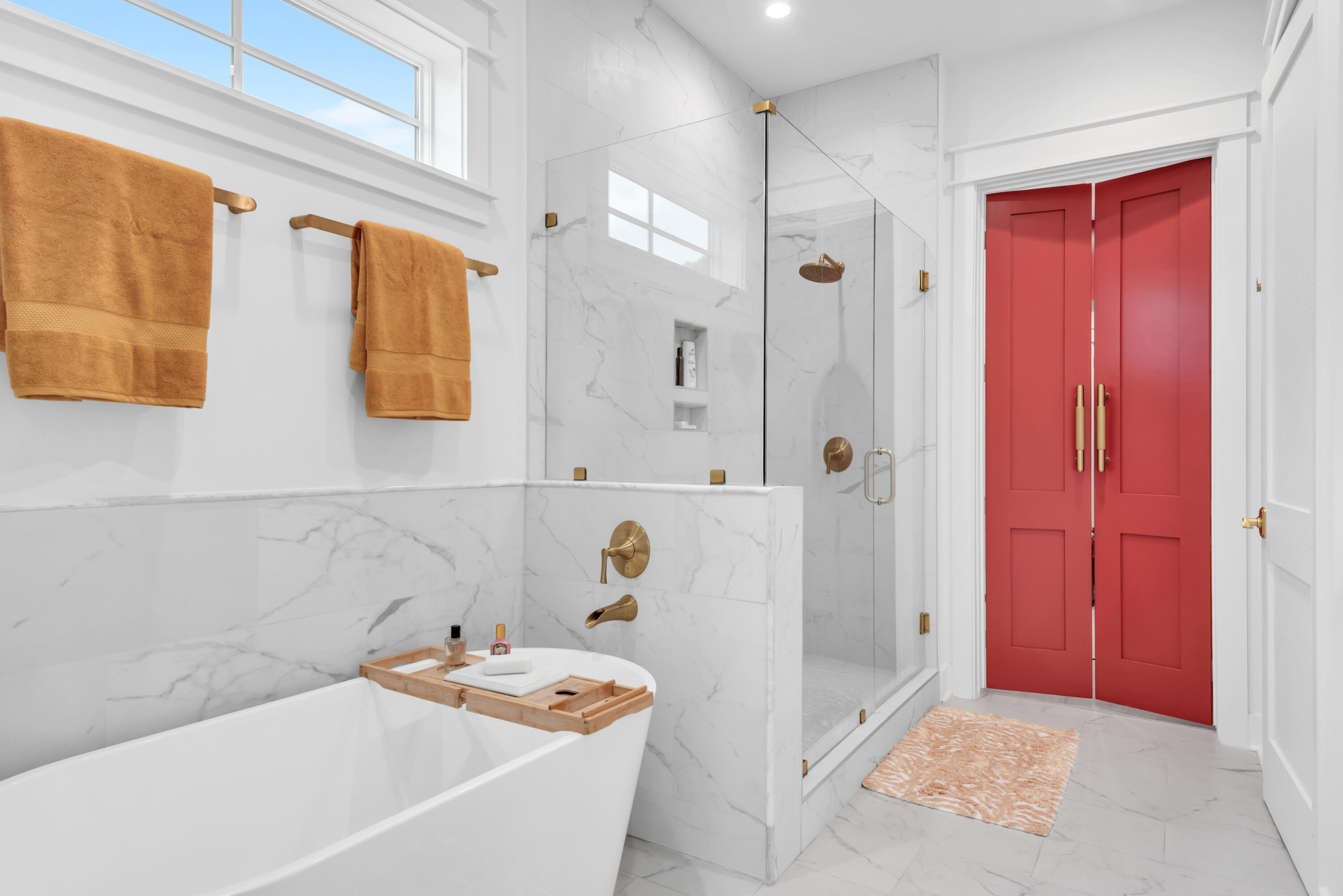 Bathroom with a freestanding tub, marble-look tile, glass shower, brass fixtures, and red double doors.