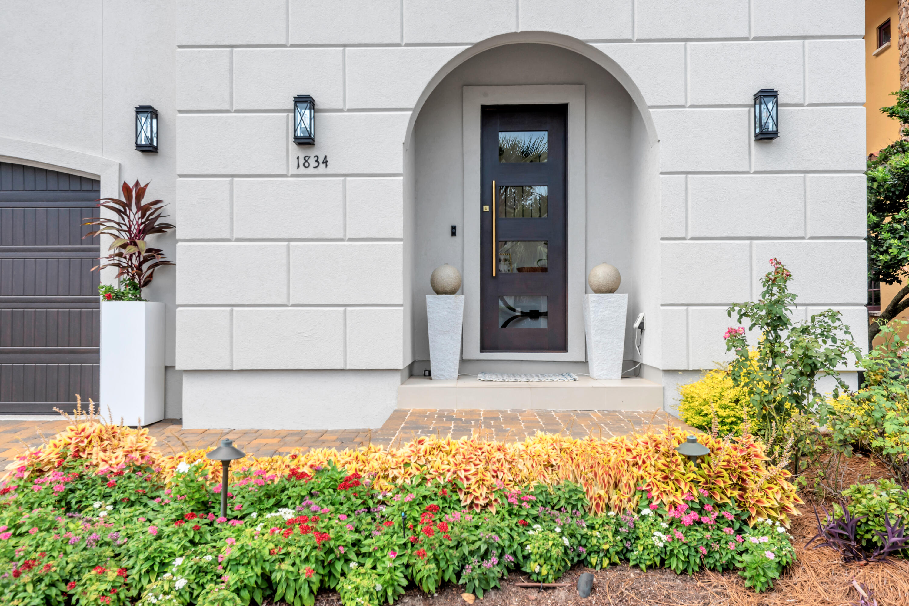 Home entrance with an arched doorway, dark glass-paneled door, and colorful flower beds.