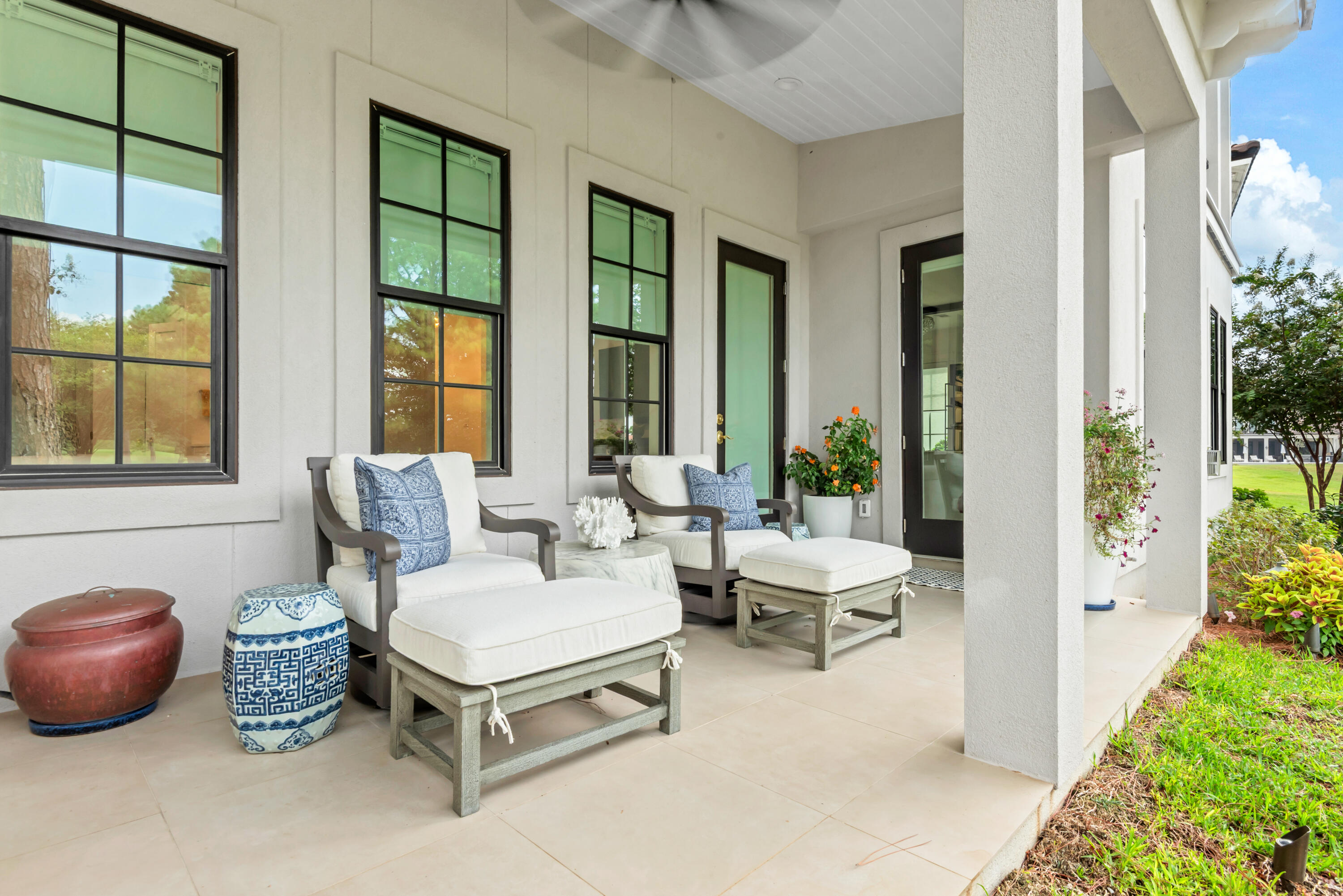 Covered porch with white-cushioned seating, blue patterned pillows, potted flowers, and white columns.