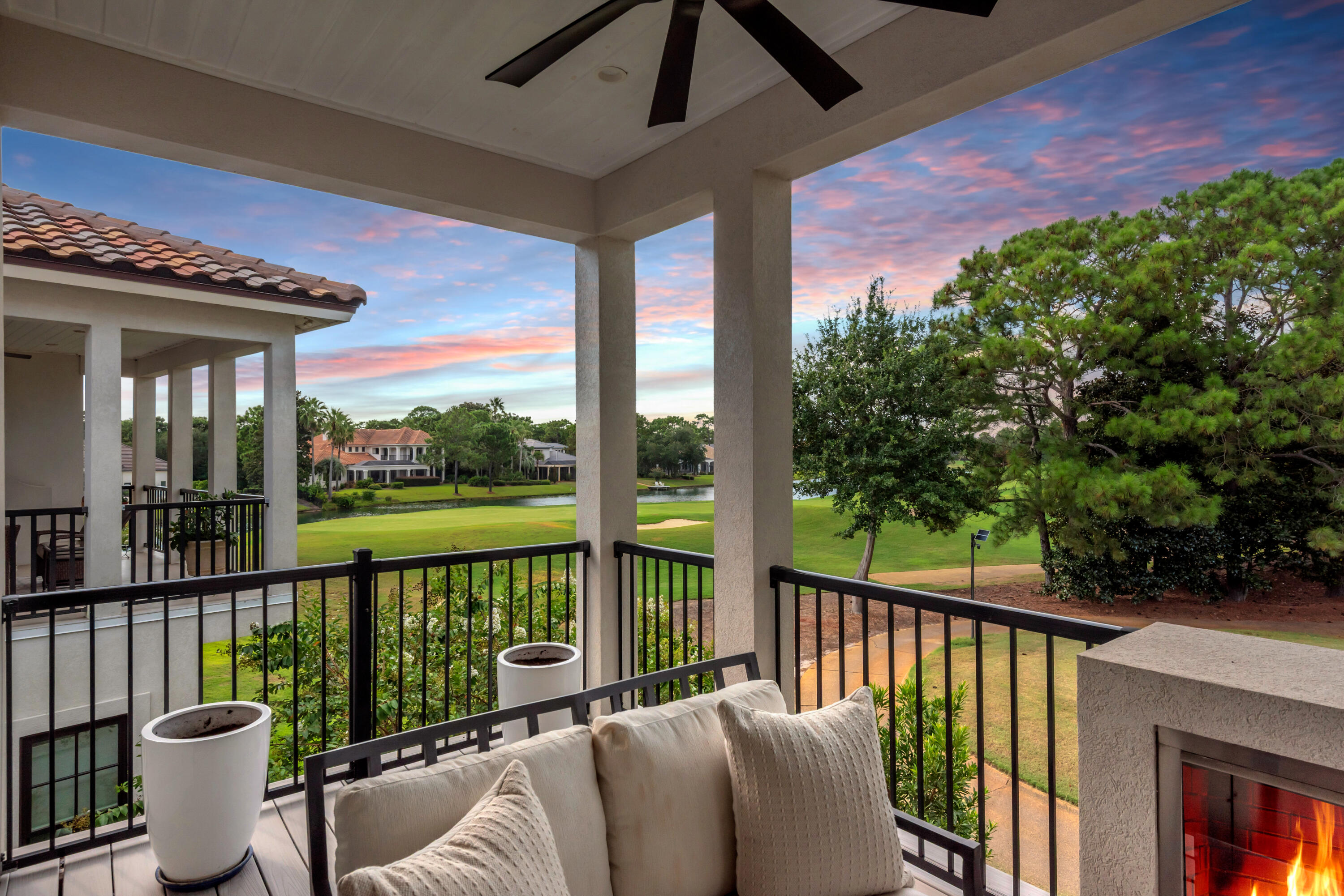 Covered balcony with cushioned seating and fireplace overlooking a golf course and water at sunset.