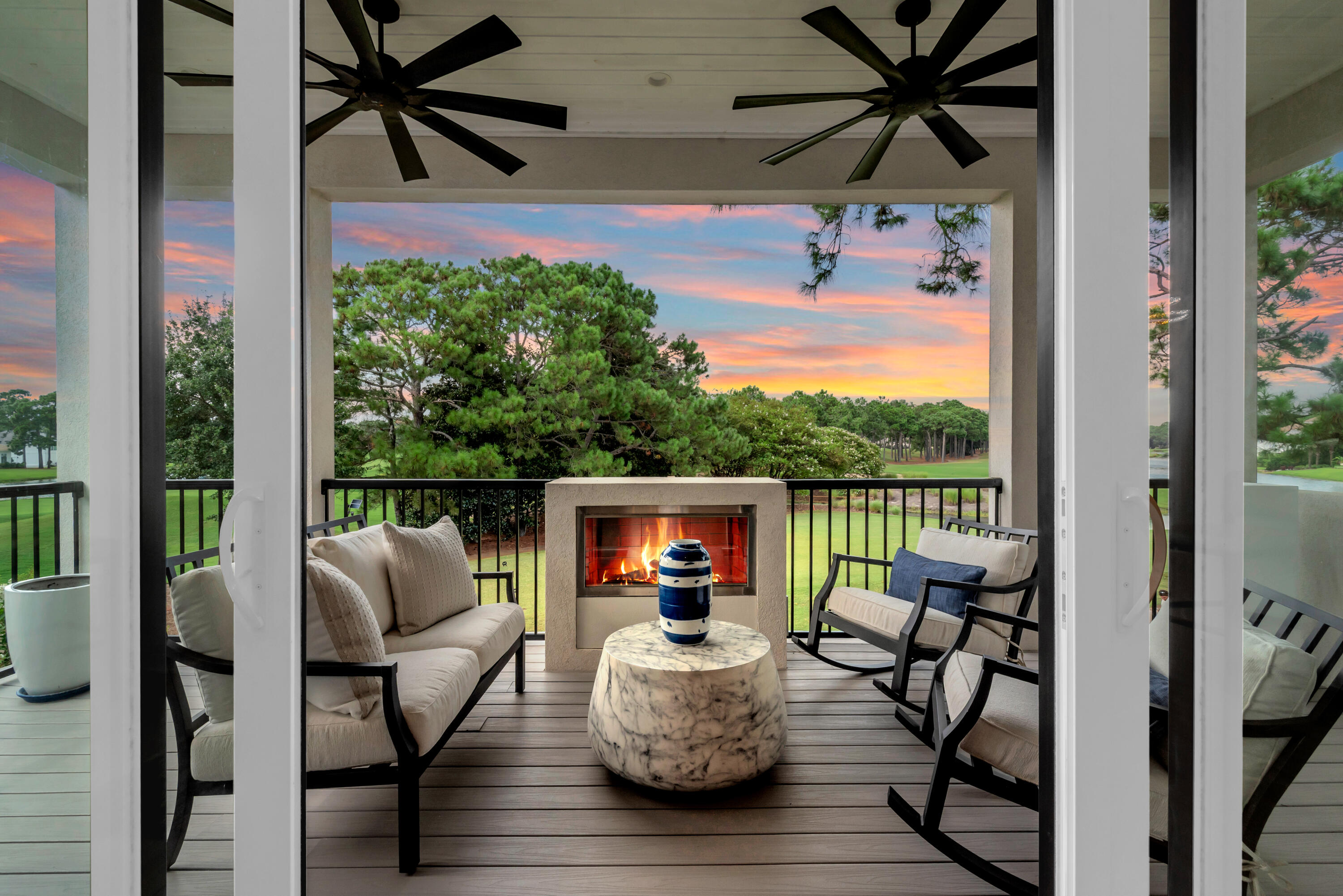 Covered balcony with a lit fireplace, cushioned seating, and a golf course view at sunset.