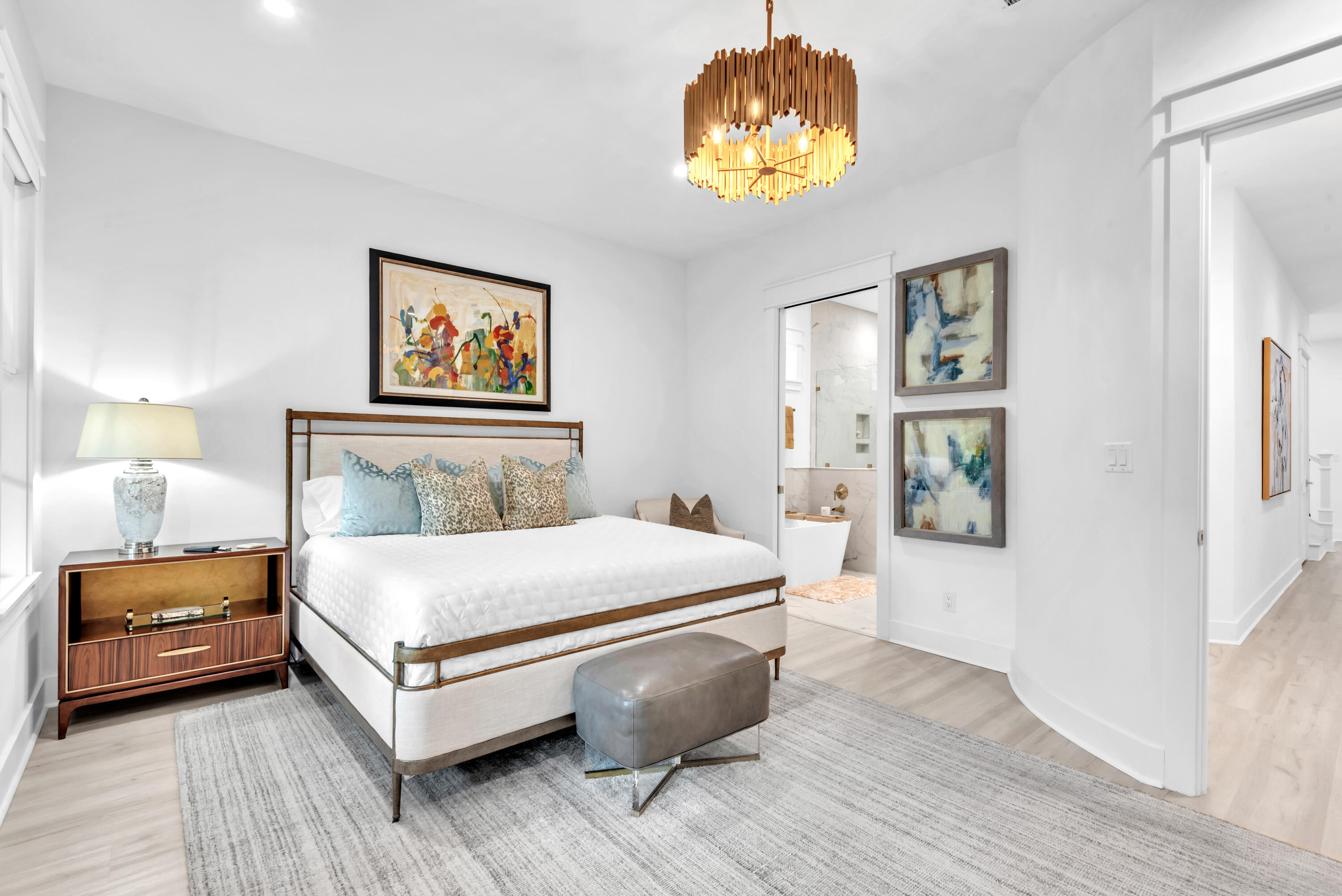 Bedroom with a white bed, gold chandelier, abstract art, and an open doorway to the bathroom.