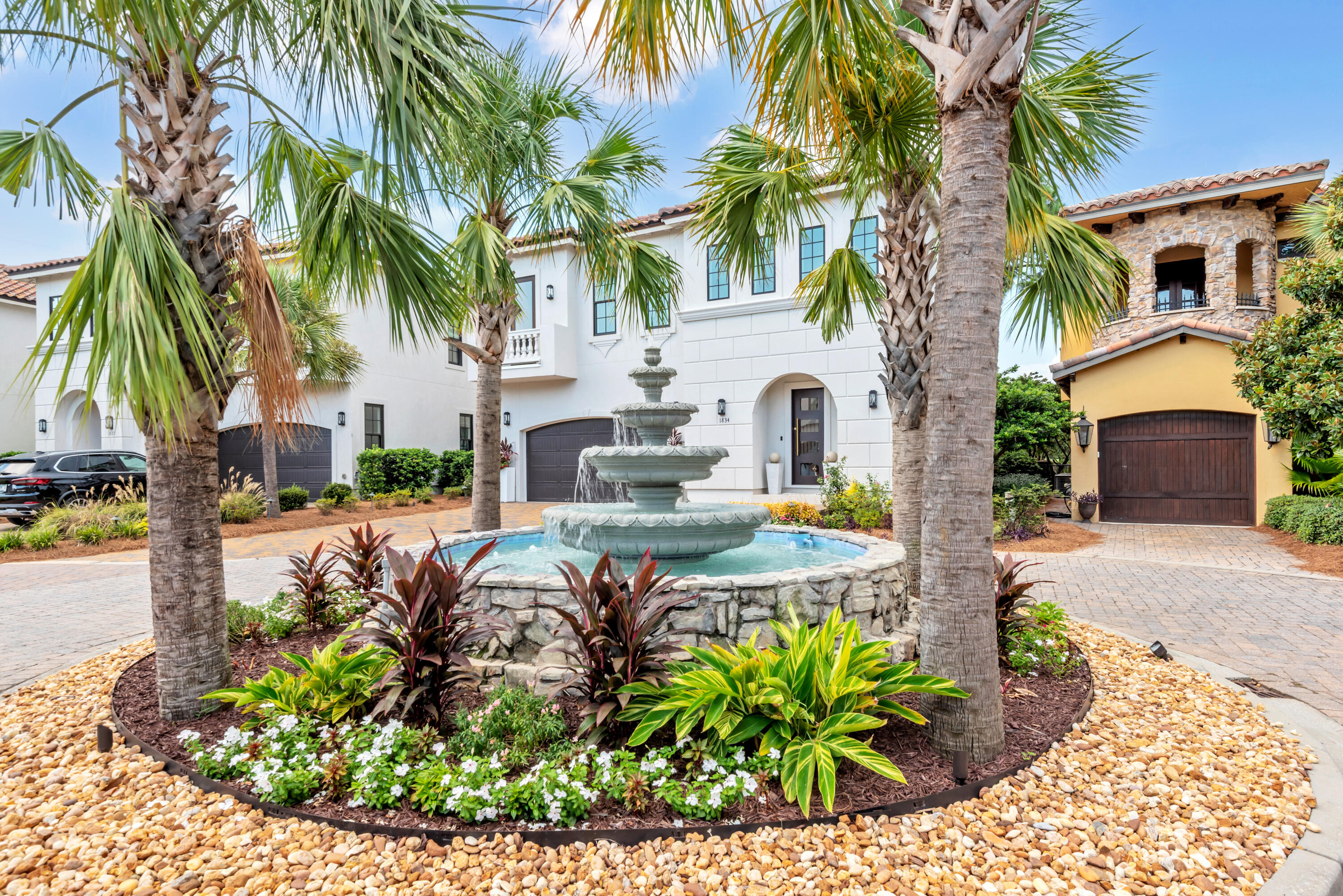 White two-story home framed by palms, with a tiered fountain and tropical landscaping in front.