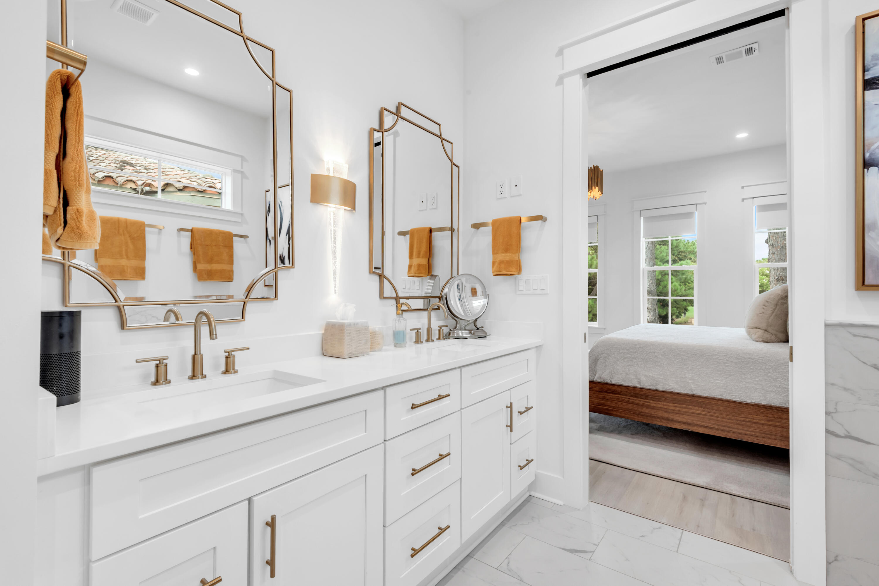 Bright white double-sink bathroom vanity with brass fixtures and a bedroom visible through the doorway.