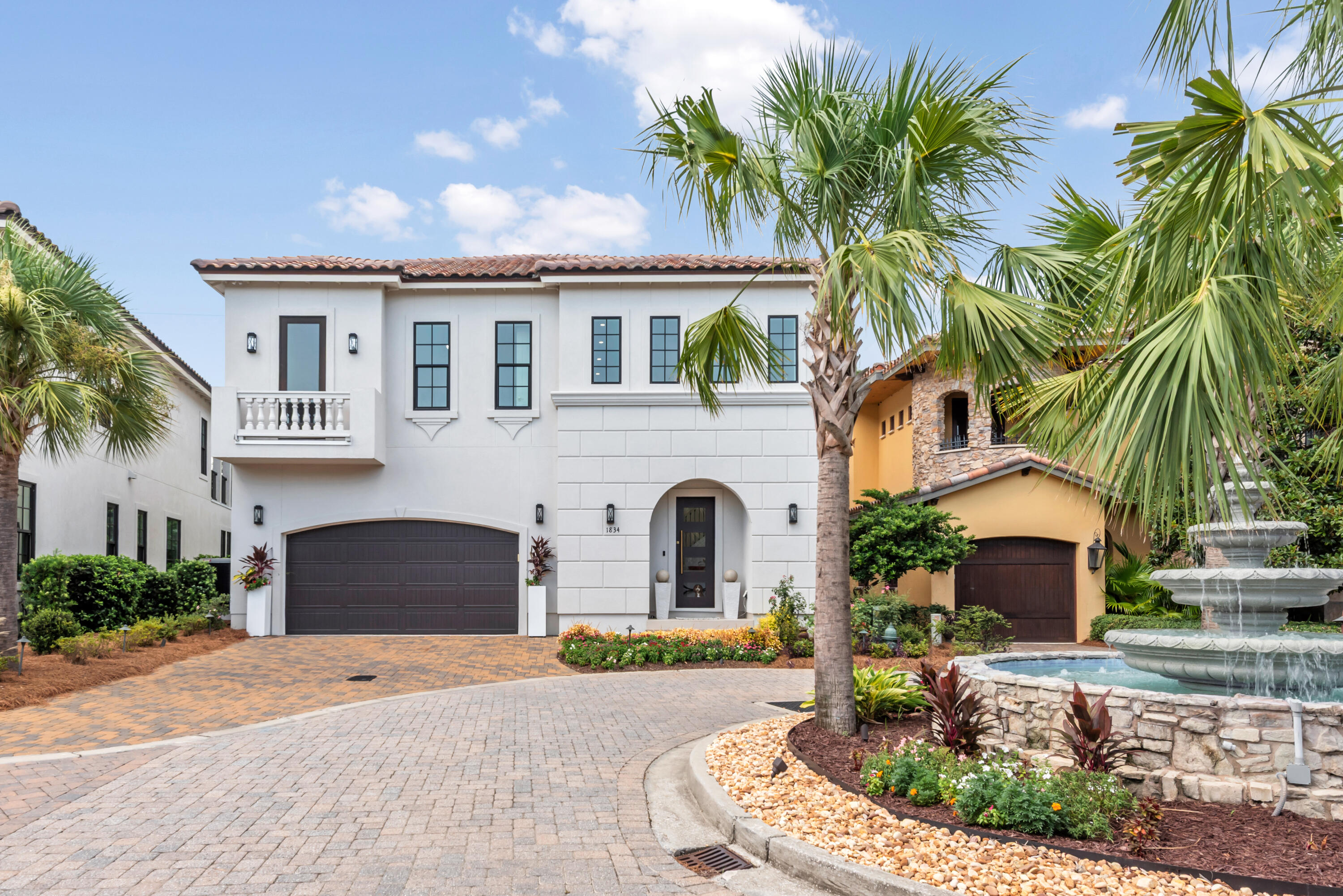 Two-story white stucco home with an arched entry, dark garage door, palms, and a stone fountain.