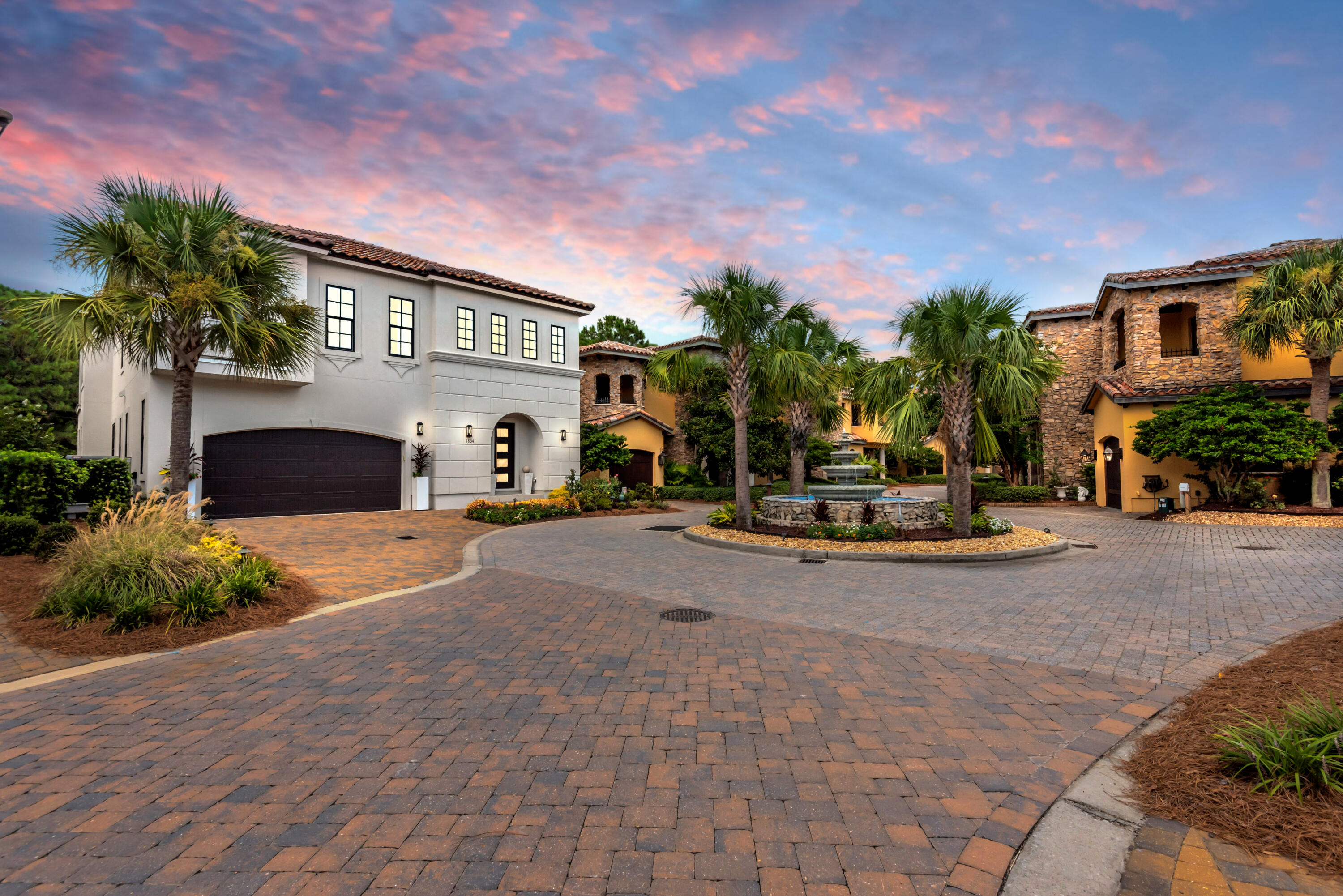 White two-story Mediterranean-style home beside a palm-lined courtyard and central fountain at dusk.
