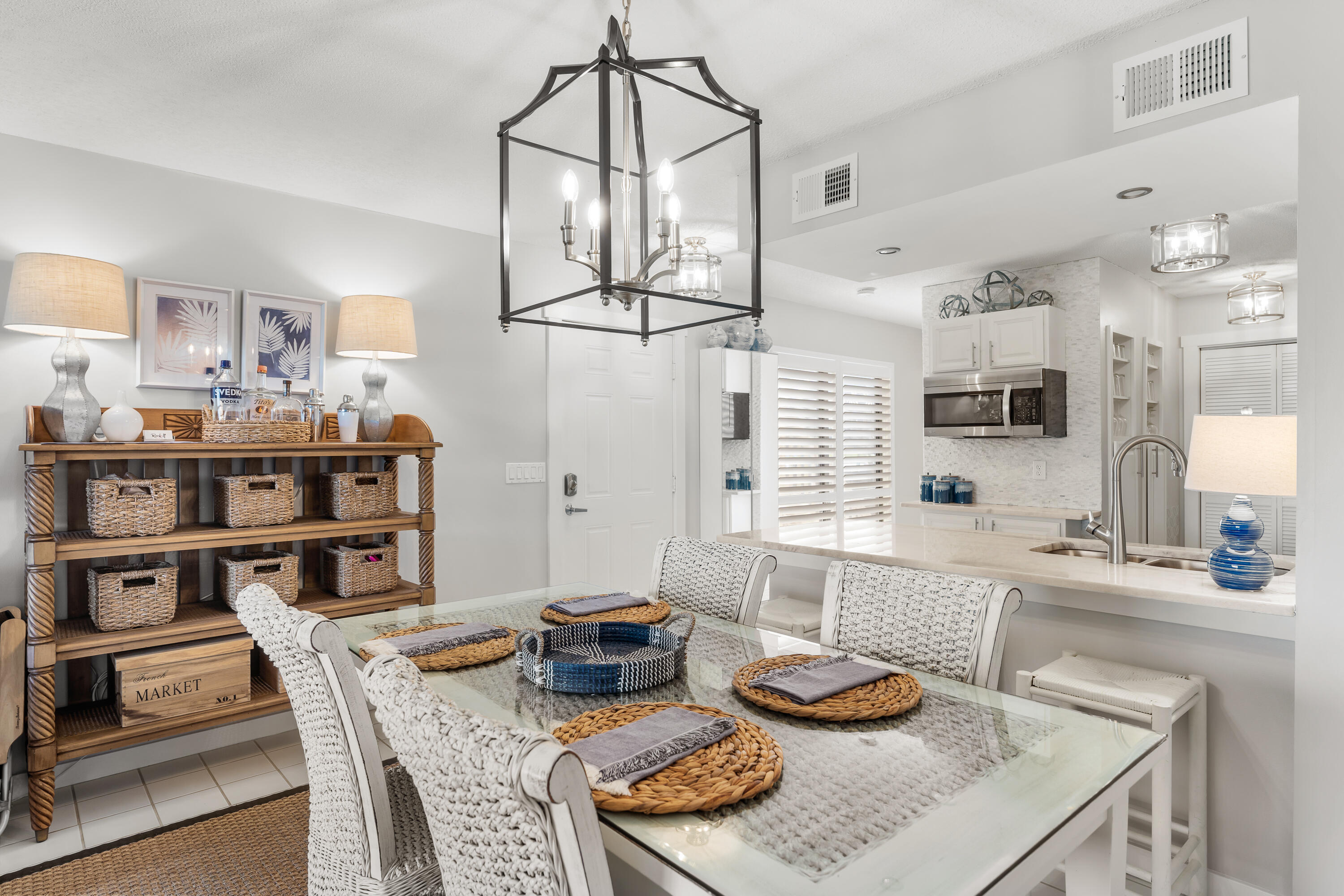 Dining area with a glass table, white woven chairs, and a large lantern pendant beside the kitchen island.