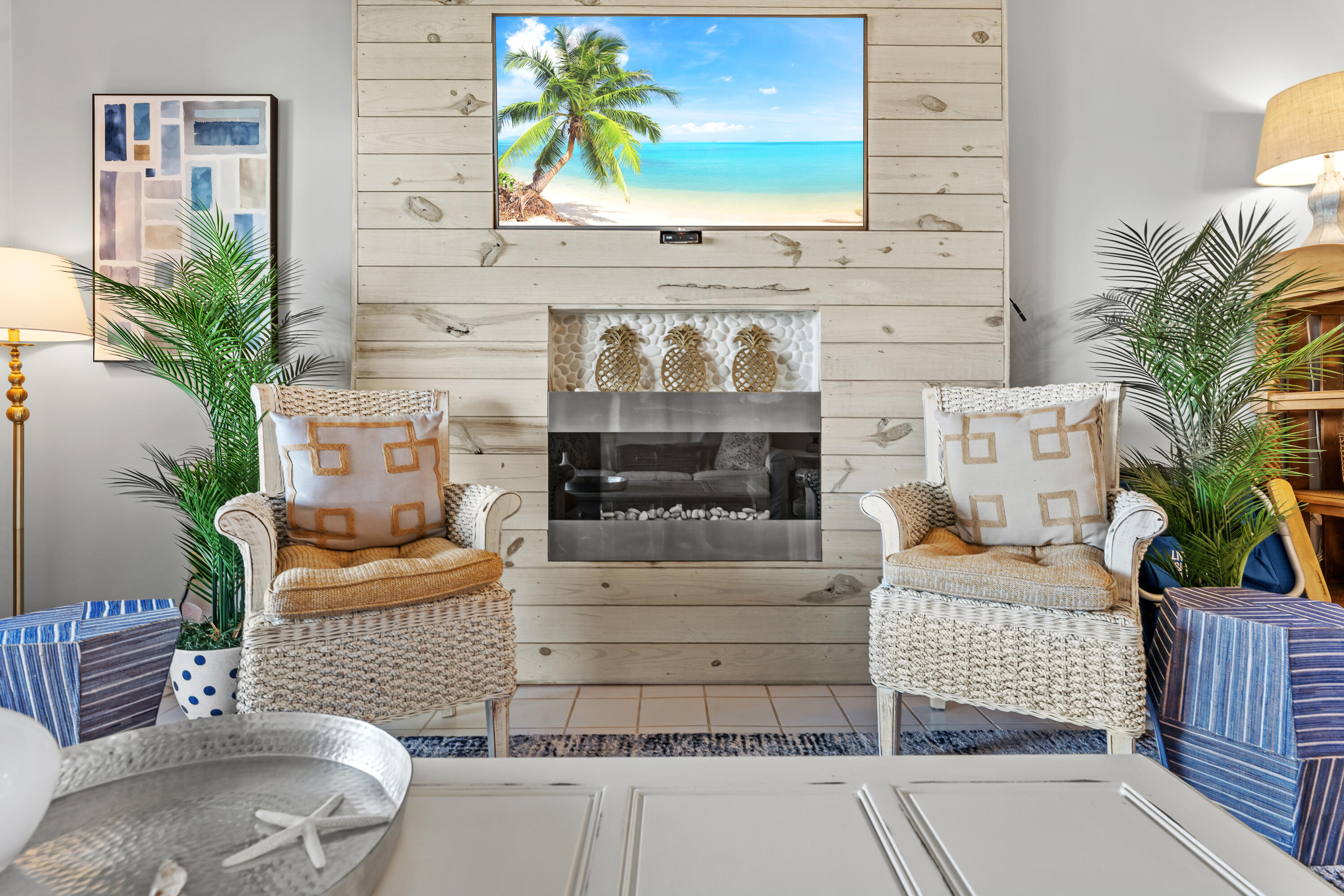 Living room with a whitewashed fireplace wall, woven armchairs, and palms.