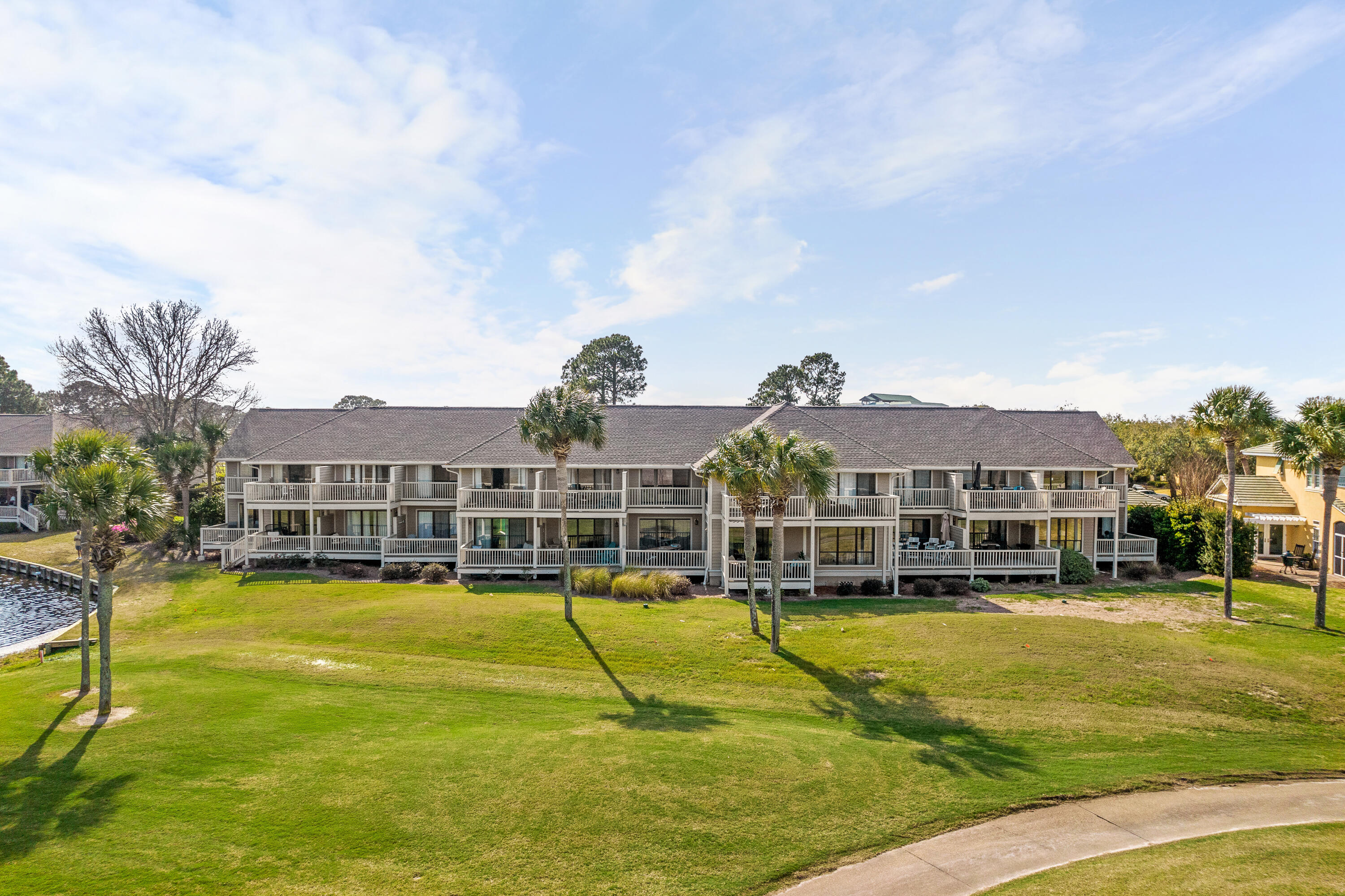 Low-rise condominium with rows of balconies, palm trees, and a lawn beside a waterway.