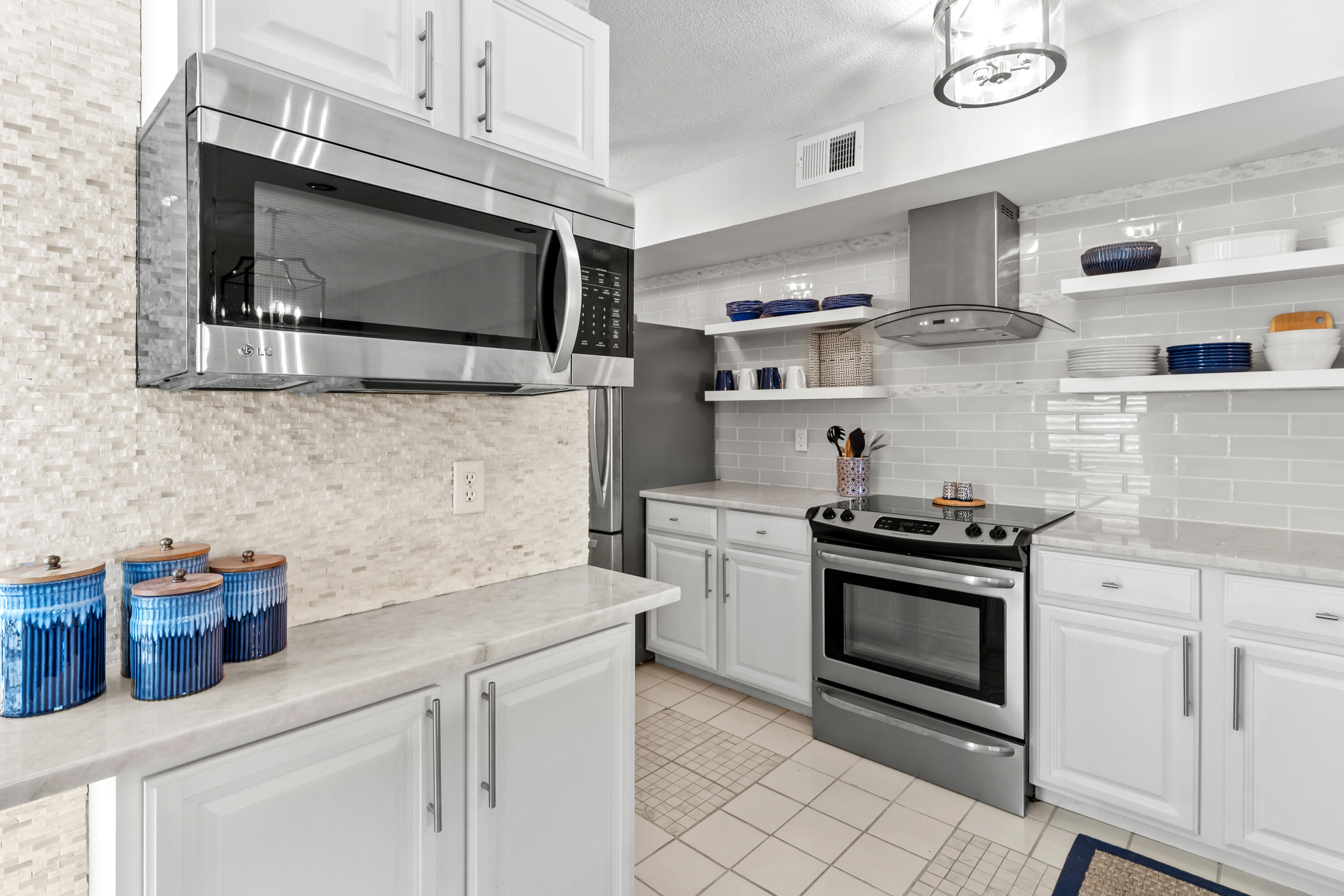 Bright kitchen with white cabinets, stainless steel appliances, glossy tile backsplash, and blue dishware on open shelves.