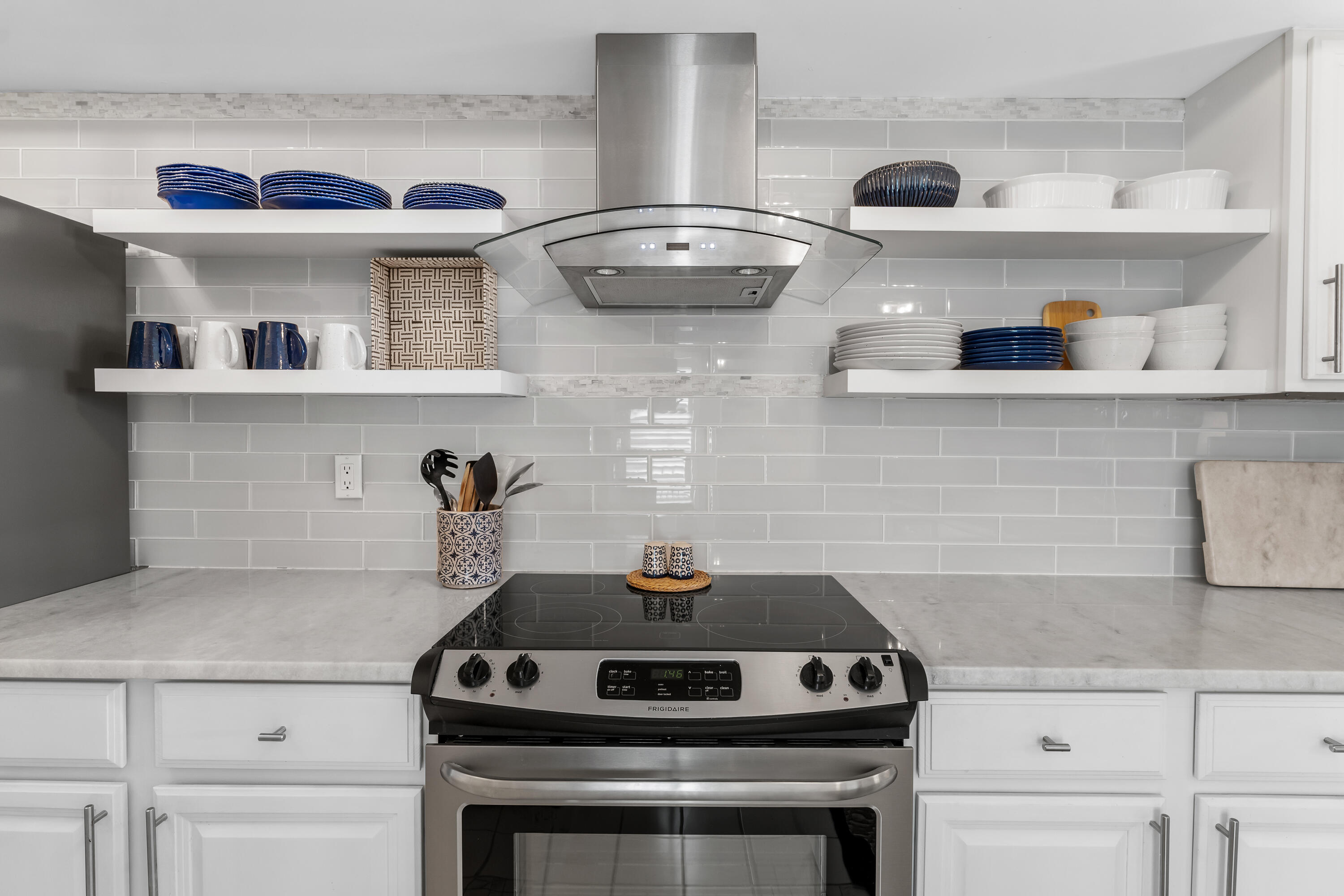 Kitchen range and oven beneath a stainless steel hood, framed by open dish shelves and a gray tile backsplash.