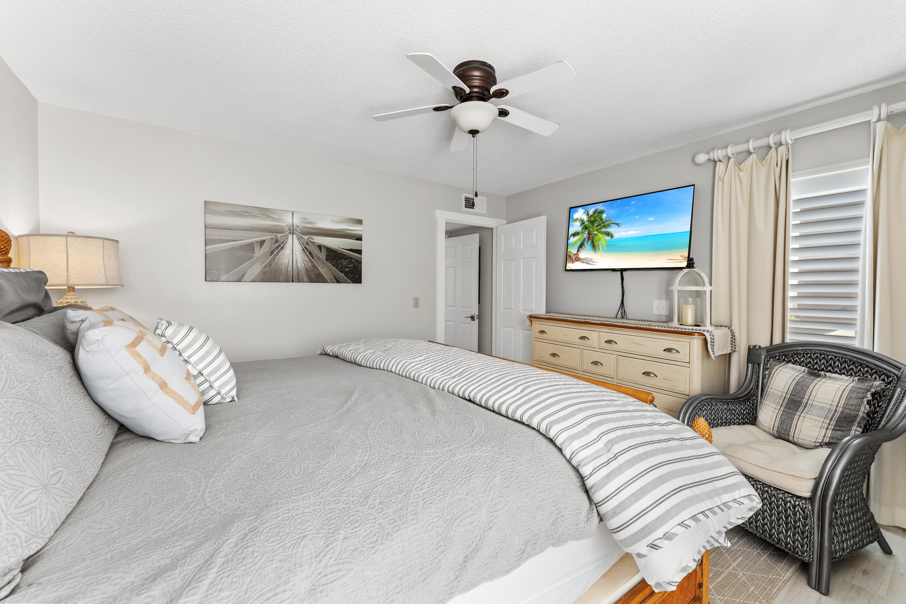 Furnished bedroom with a large gray bed, woven armchair, light wood dresser, and beach scene on the television.