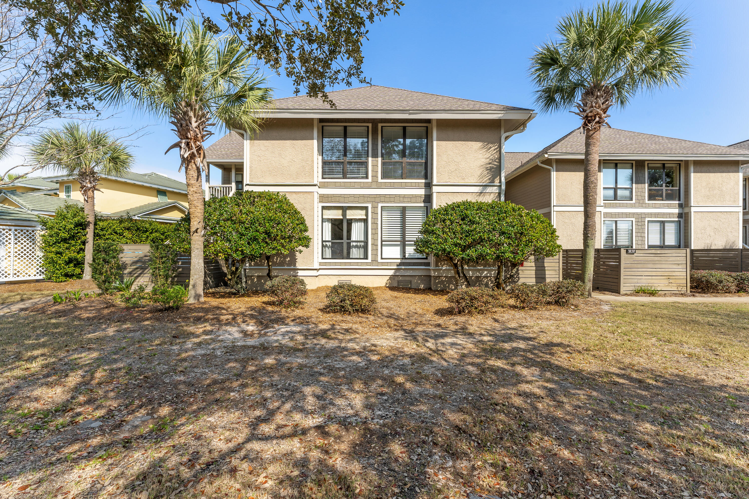 Two-story beige townhouse with palm trees, trimmed shrubs, and fenced patio areas.