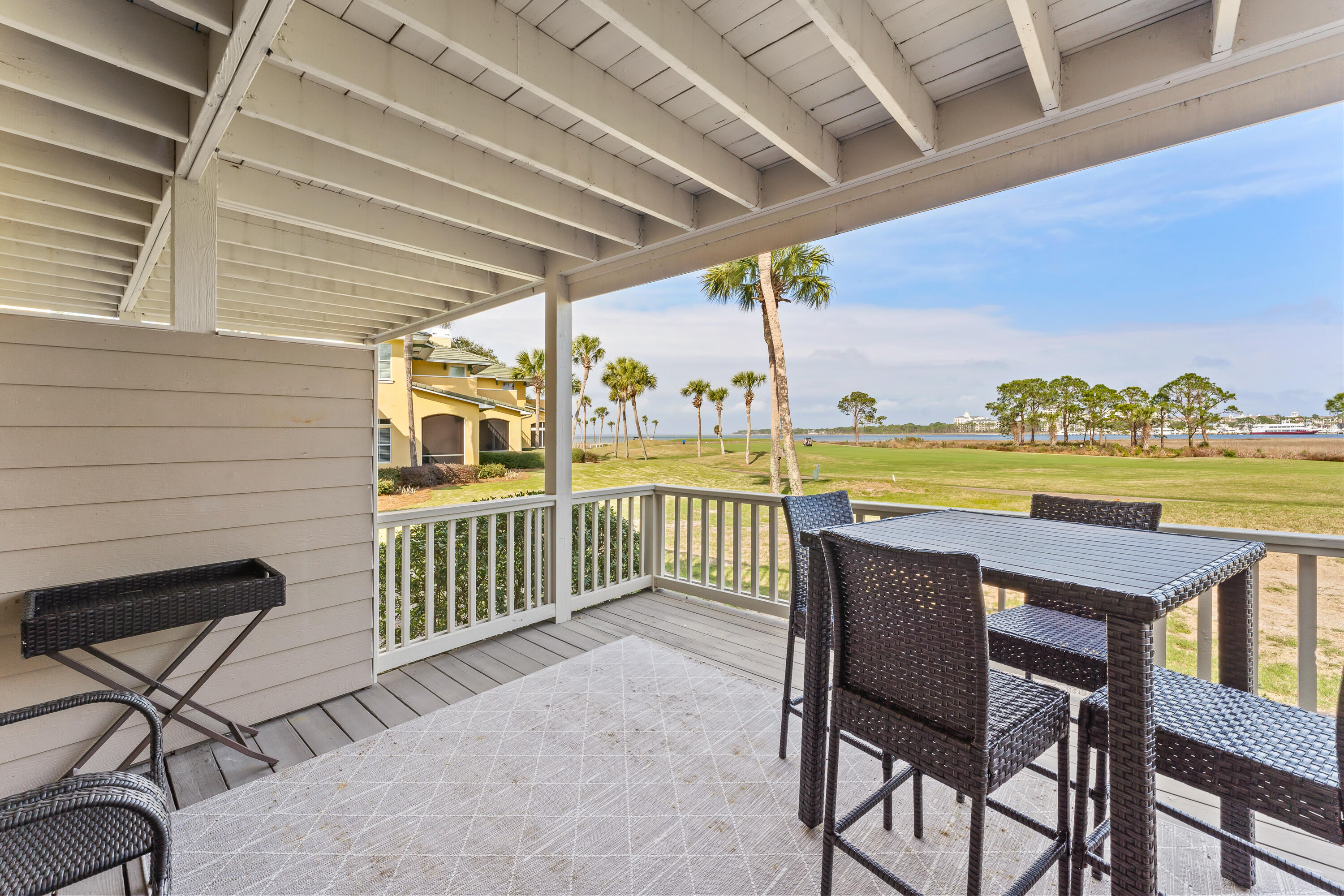 Covered patio with wicker dining set overlooking a palm-lined lawn and waterfront.