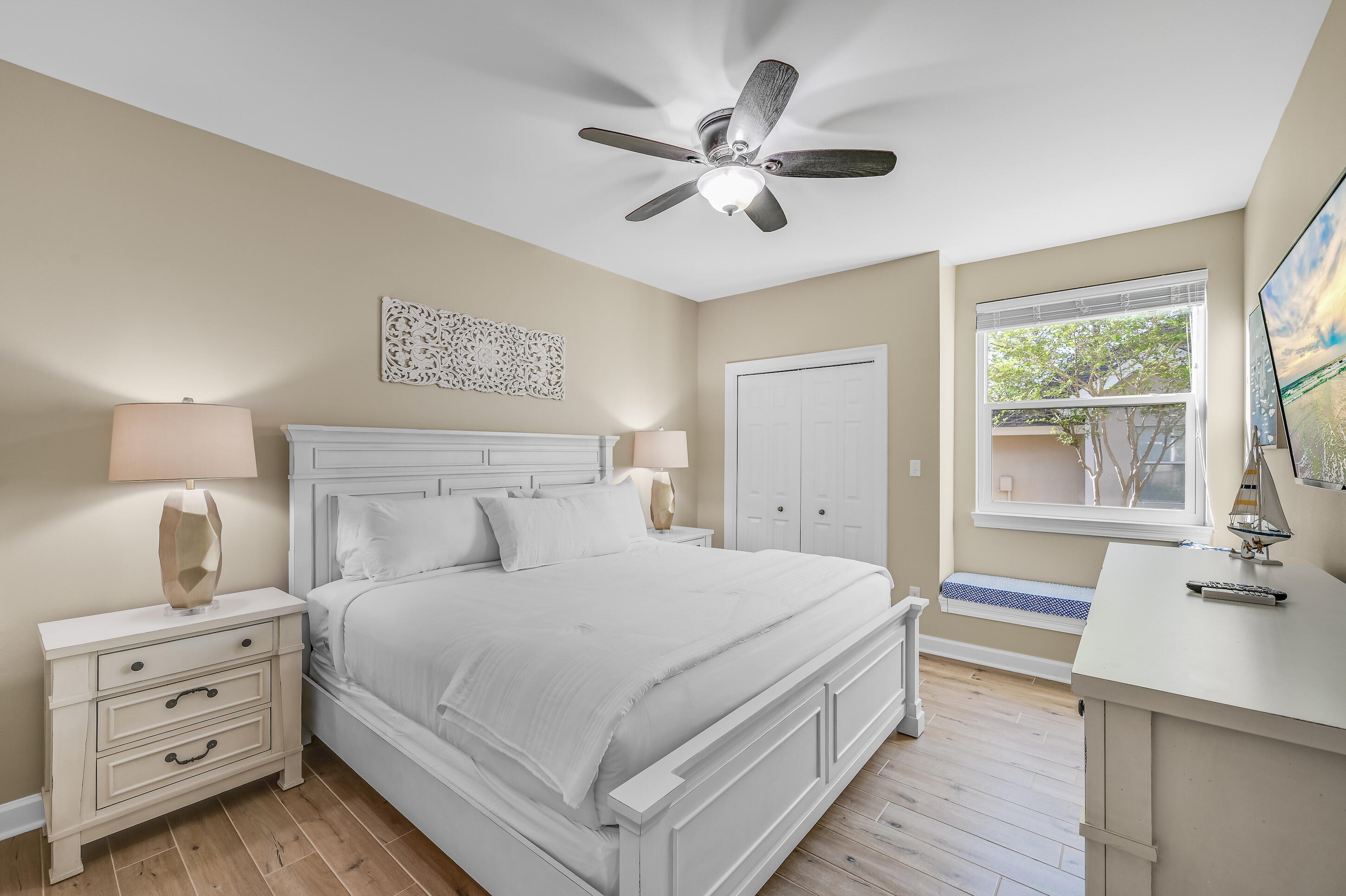 Bedroom with a white paneled bed, ceiling fan, and window overlooking trees.